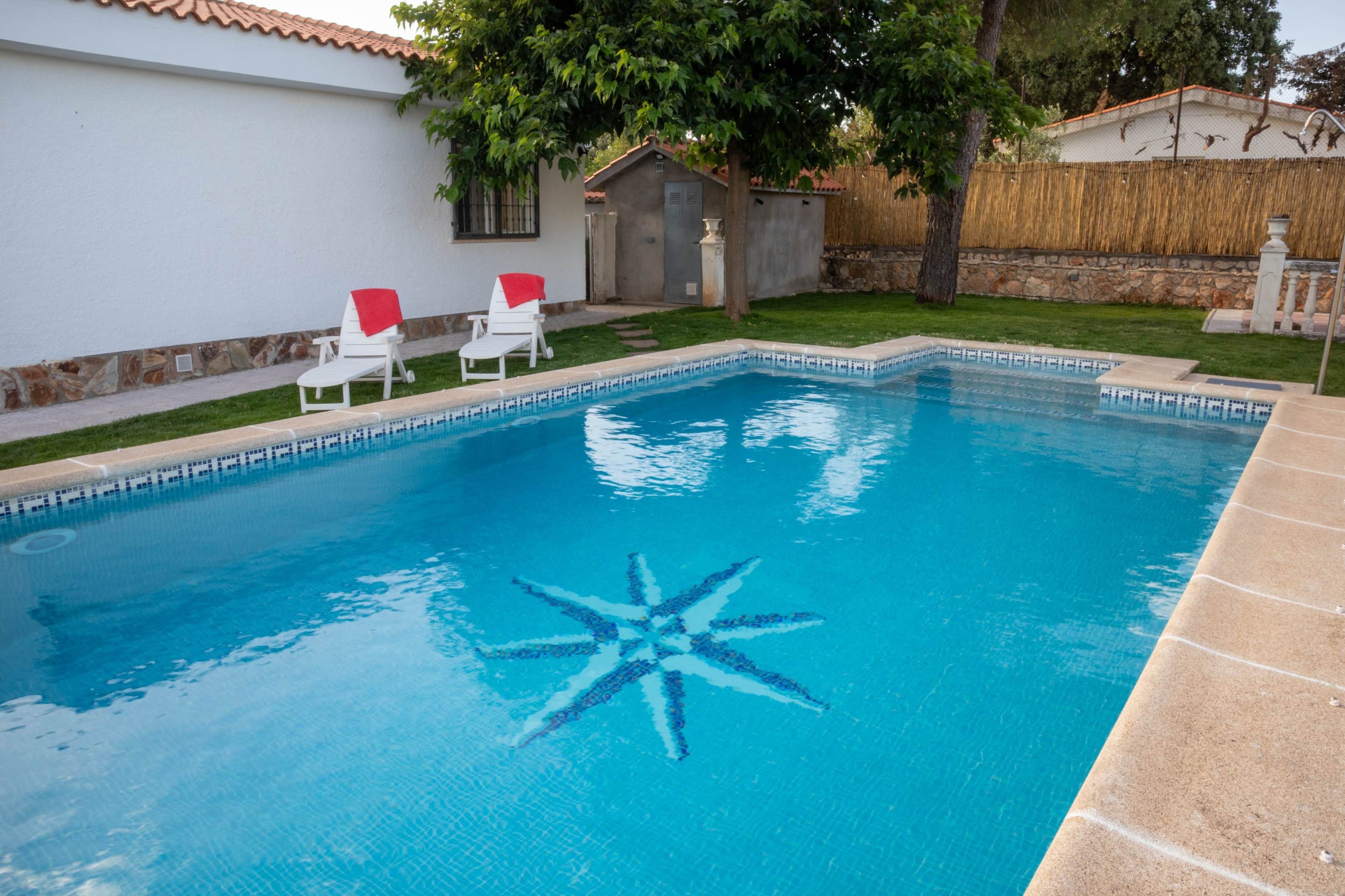 The image shows a swimming pool with a star-shaped design at the bottom, surrounded by white lounge chairs and a grassy area, with a wooden fence and trees in the background.