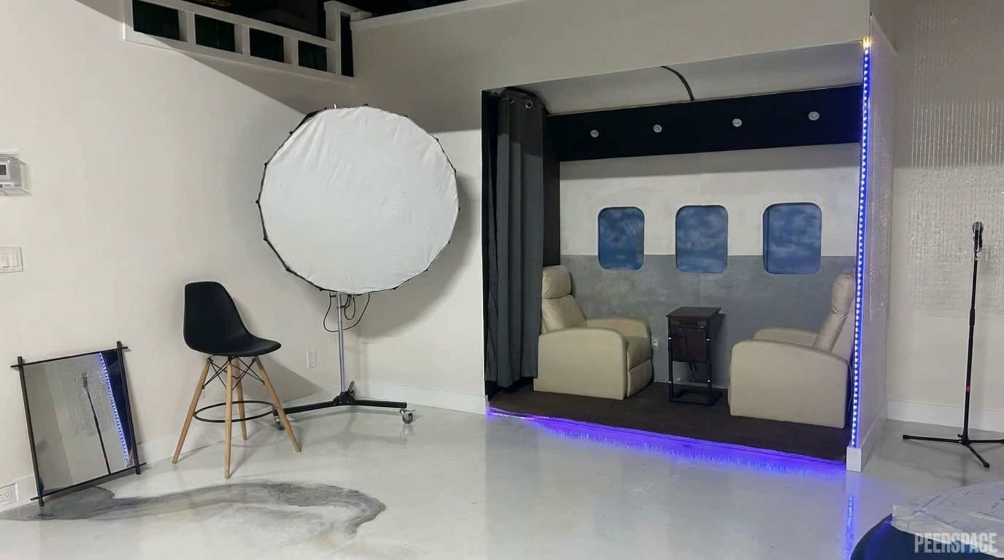 The image shows a small staged interior resembling an airplane cabin, featuring two seats and a backdrop with window graphics, alongside a photography lighting setup and a chair.