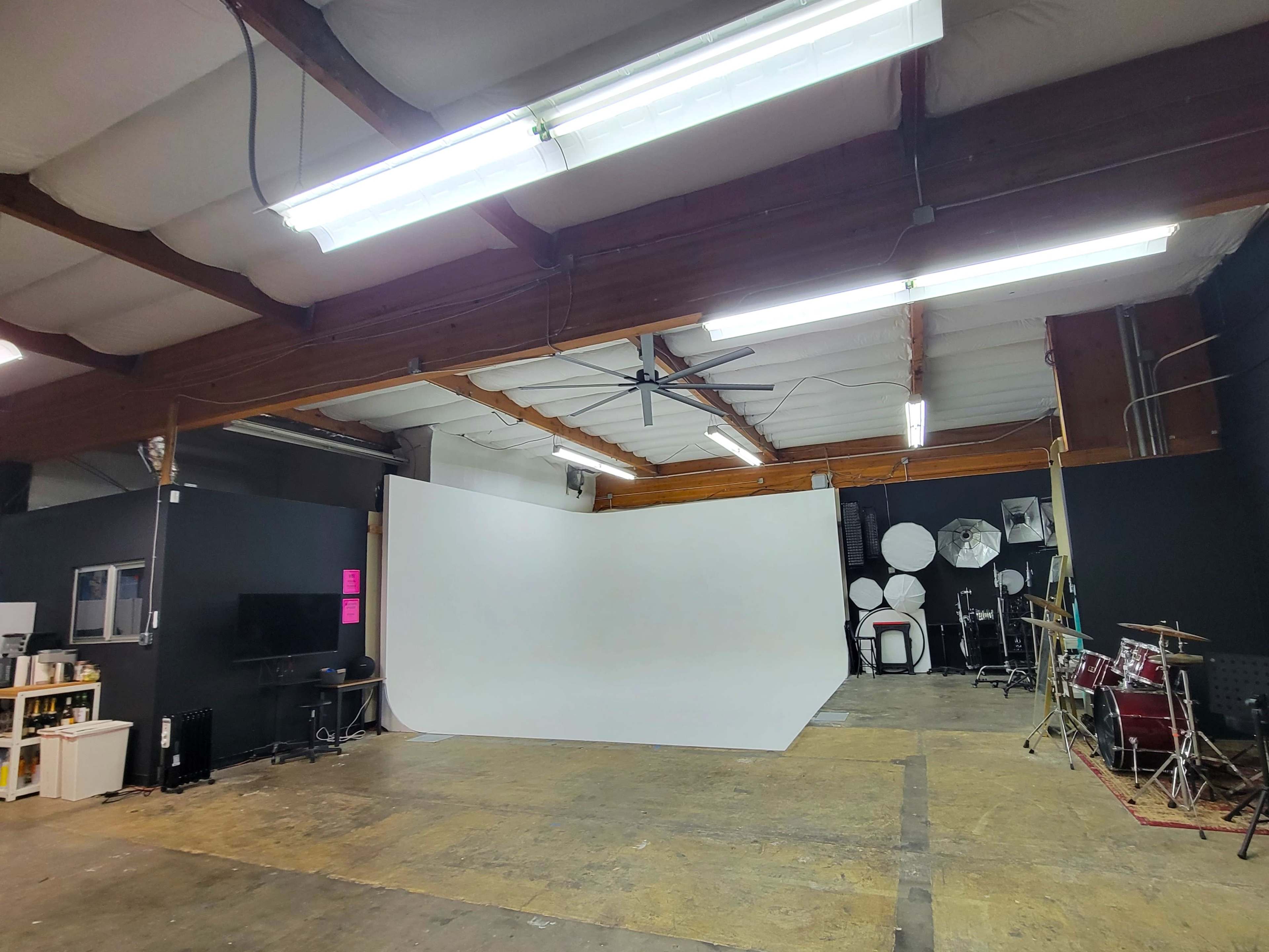 The image shows a spacious indoor studio with a white backdrop, some lighting fixtures, a television, and a drumming setup.