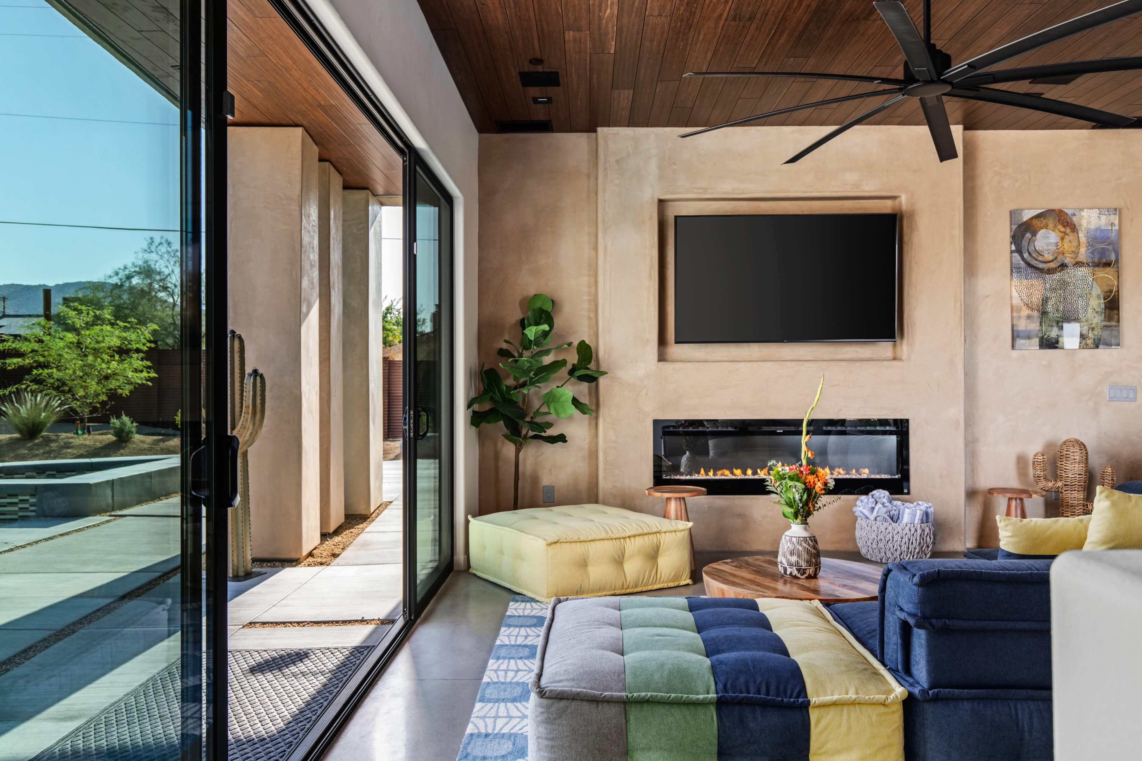 A modern living room features a large wall-mounted TV, a minimalist fireplace, and a blend of colorful seating options, with glass doors opening to a landscaped outdoor area.