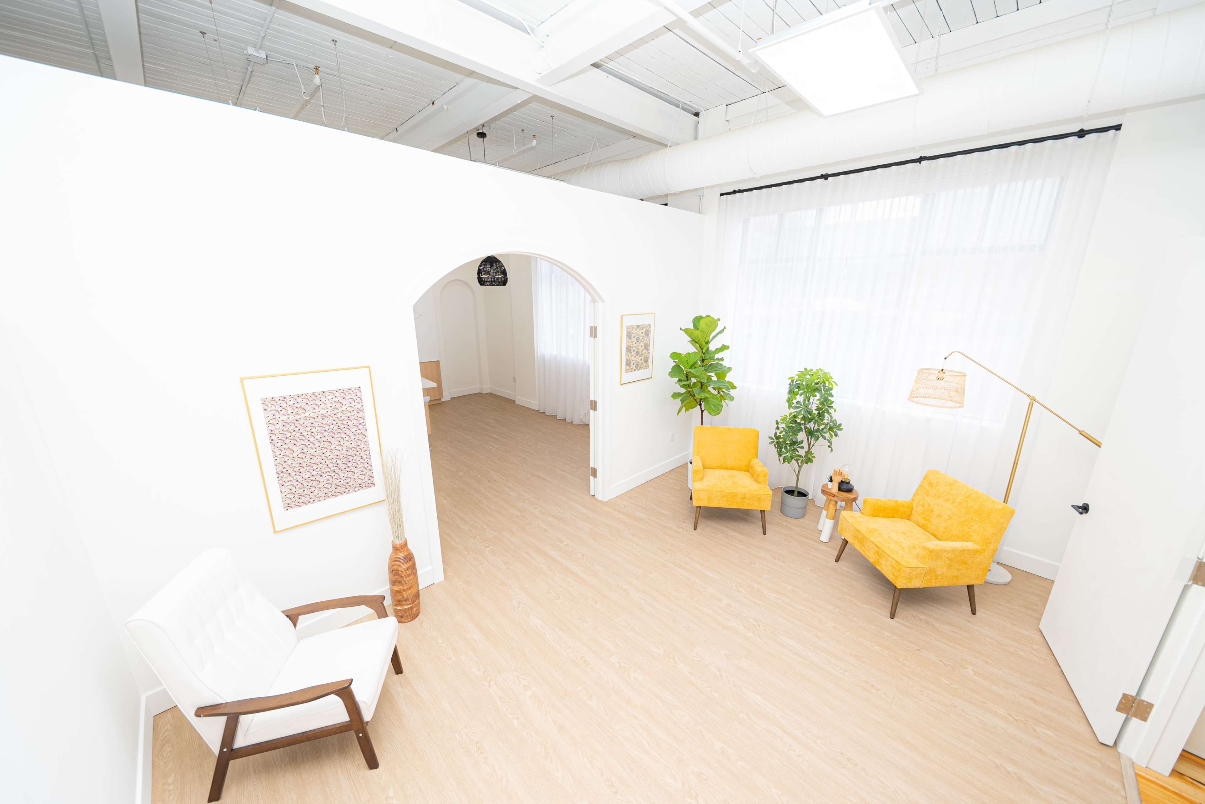A bright, minimalist room with two yellow chairs, a small table, and greenery, featuring a large window with sheer curtains.