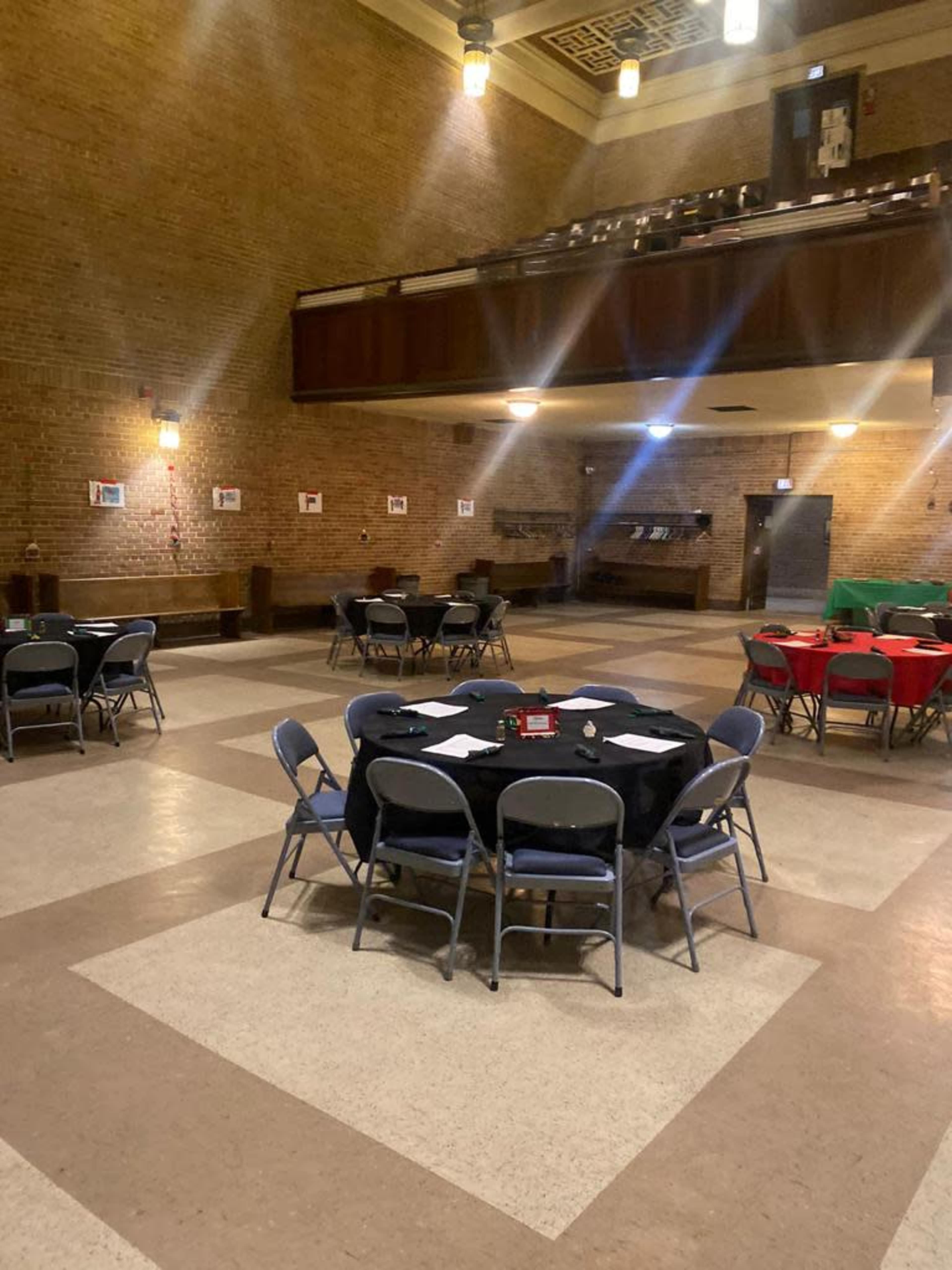 A large, empty hall features several round tables, some covered with black and red tablecloths, set against a backdrop of brick walls and wooden accents.