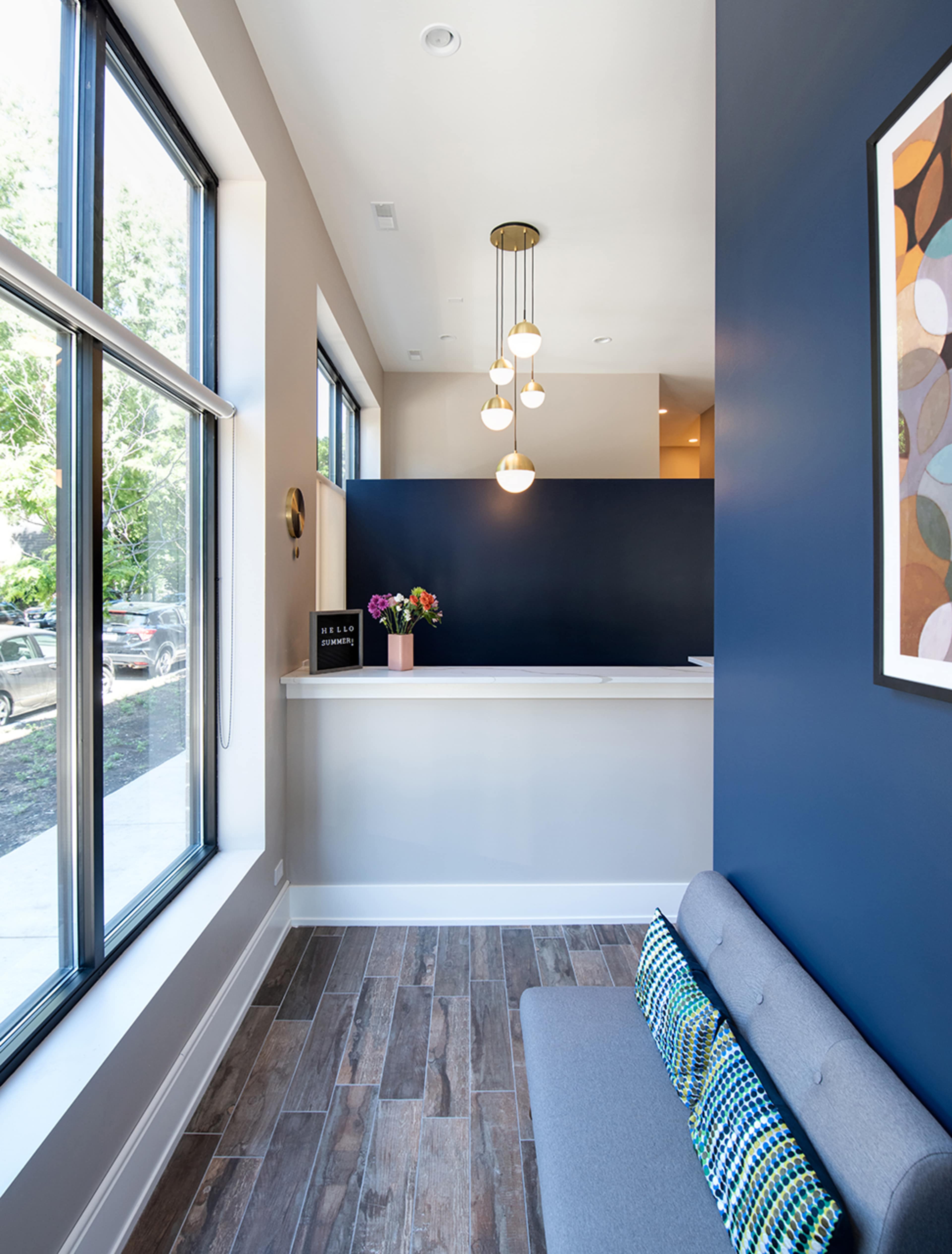 A modern reception area with a gray bench, large windows, and a blue accent wall featuring pendant lighting and a floral arrangement on the desk.