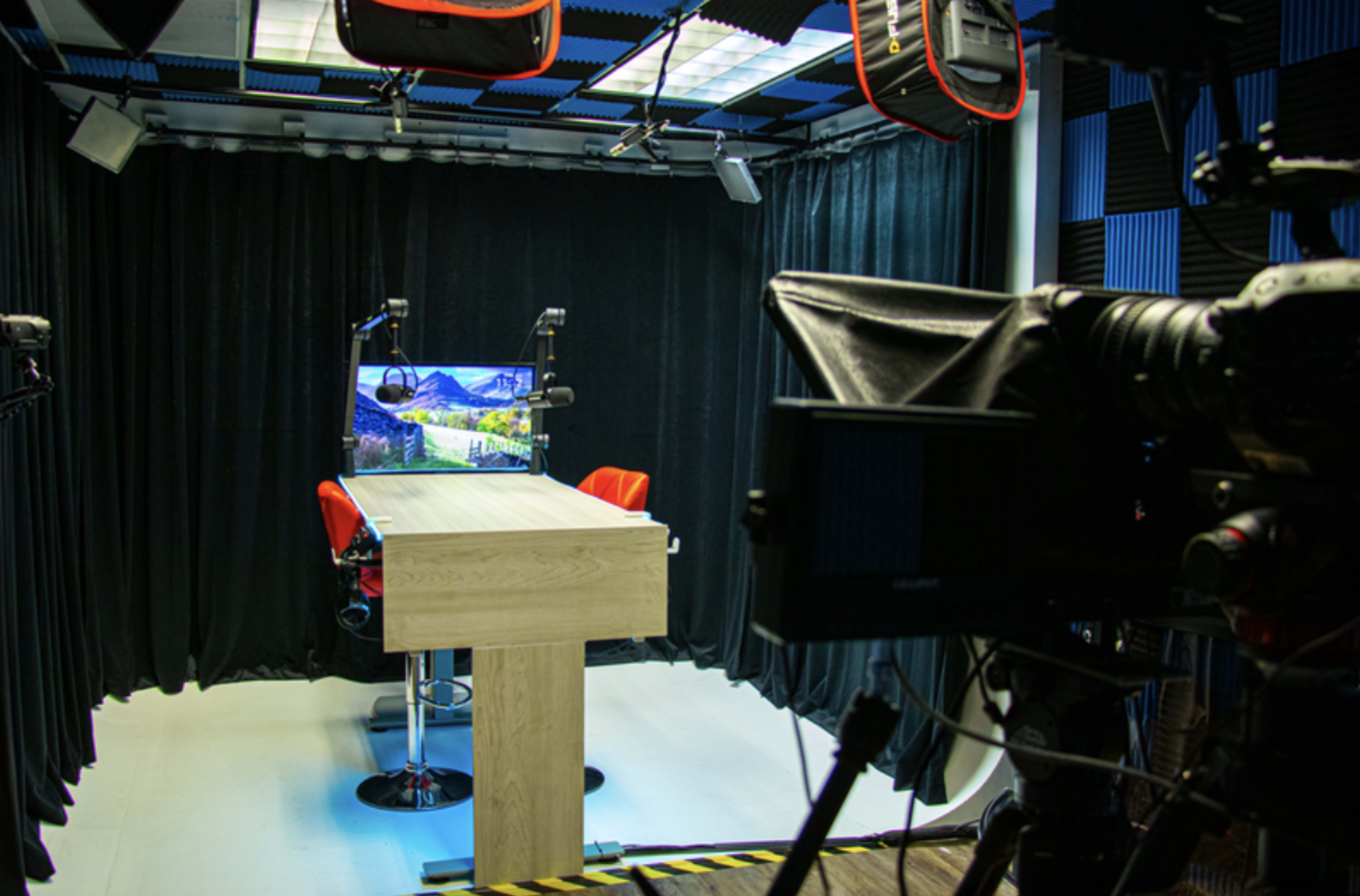 A video studio setup with a central table, two red chairs, a large monitor displaying a landscape, and various lighting equipment overhead.