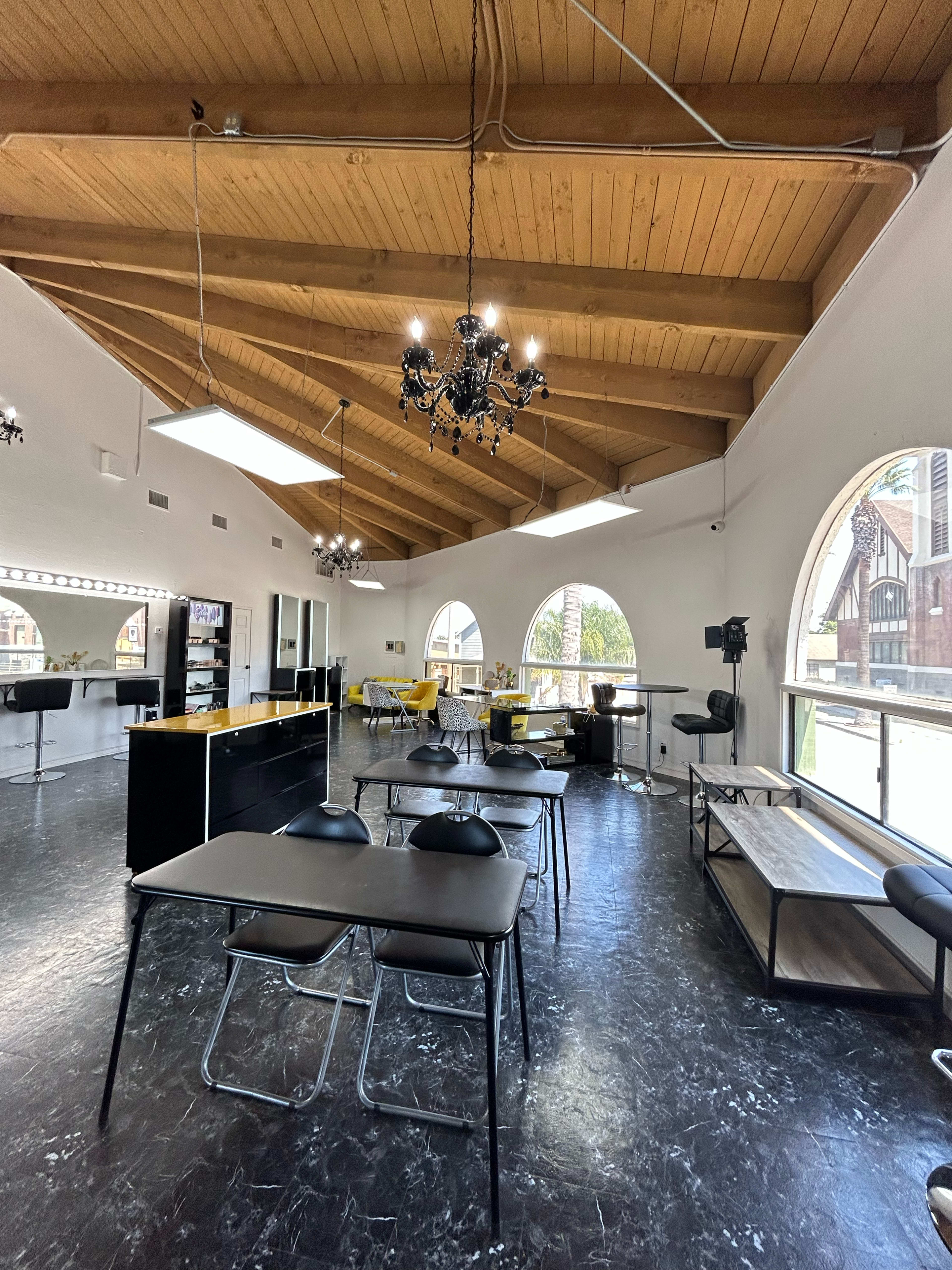 The image shows a spacious salon interior with high wooden ceilings, several tables, and large windows overlooking an outside view.