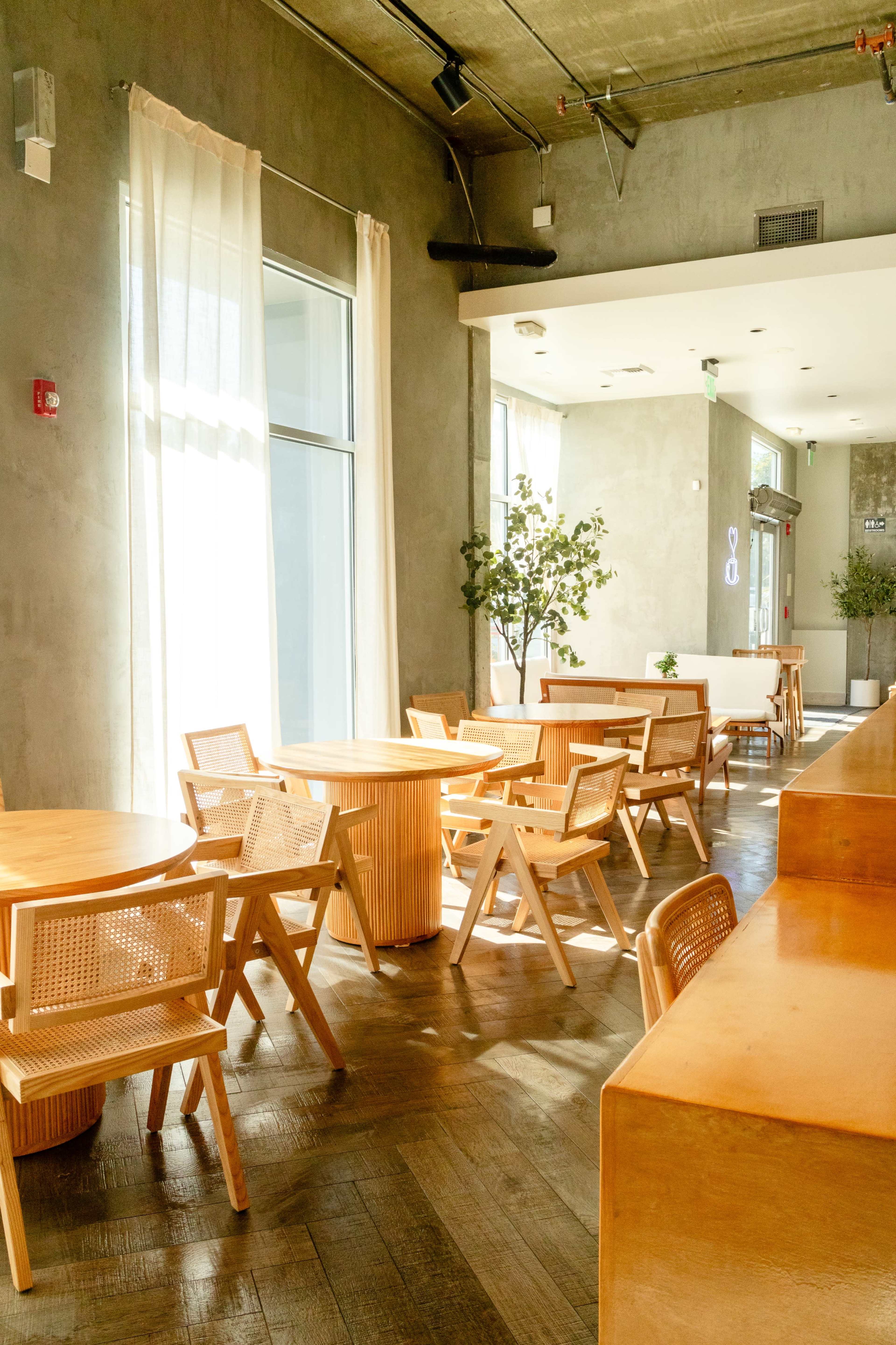 A modern, minimalist restaurant interior with wooden tables and chairs arranged neatly, illuminated by natural light.