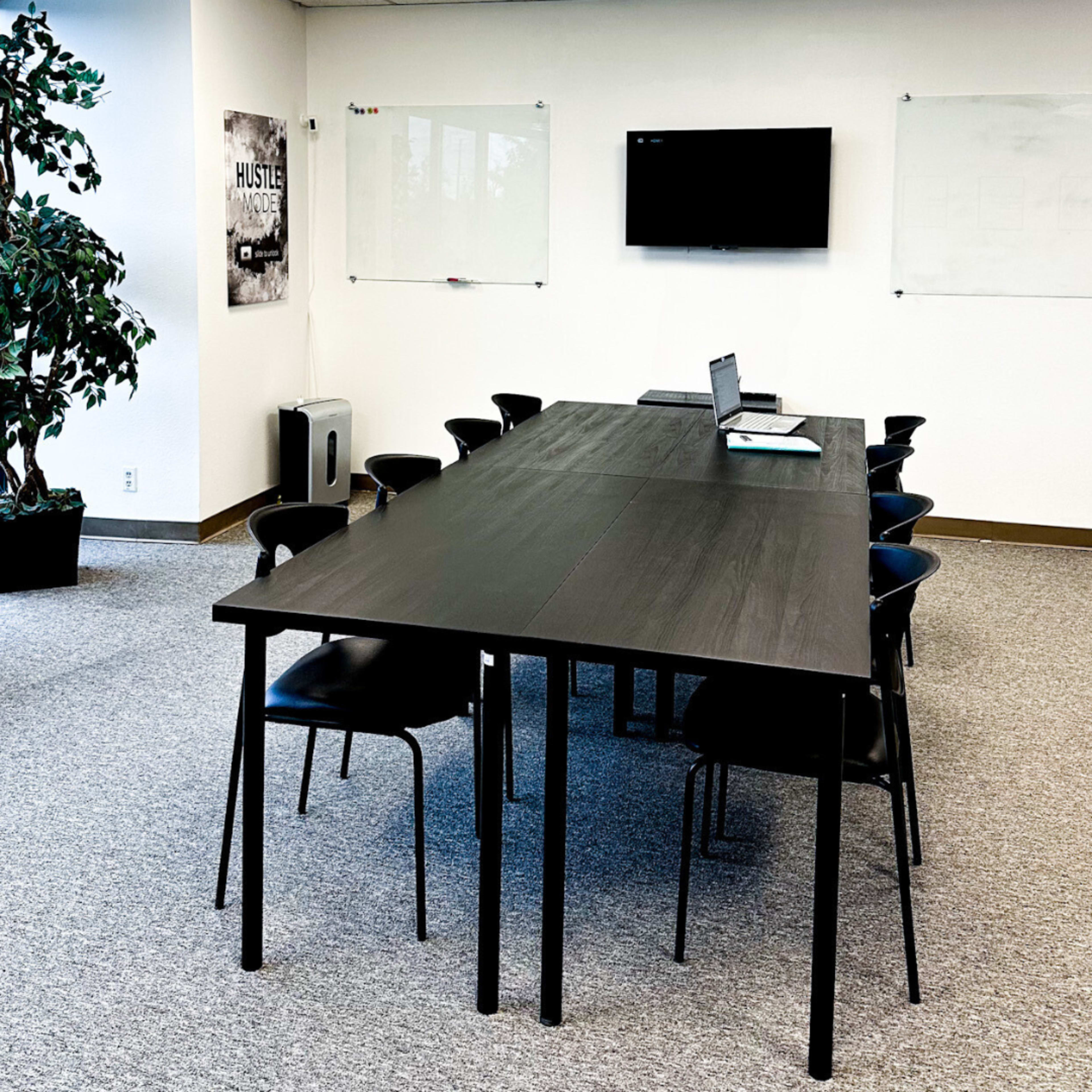 A long wooden table surrounded by black chairs is set in a meeting room with a television screen and whiteboards on the walls.