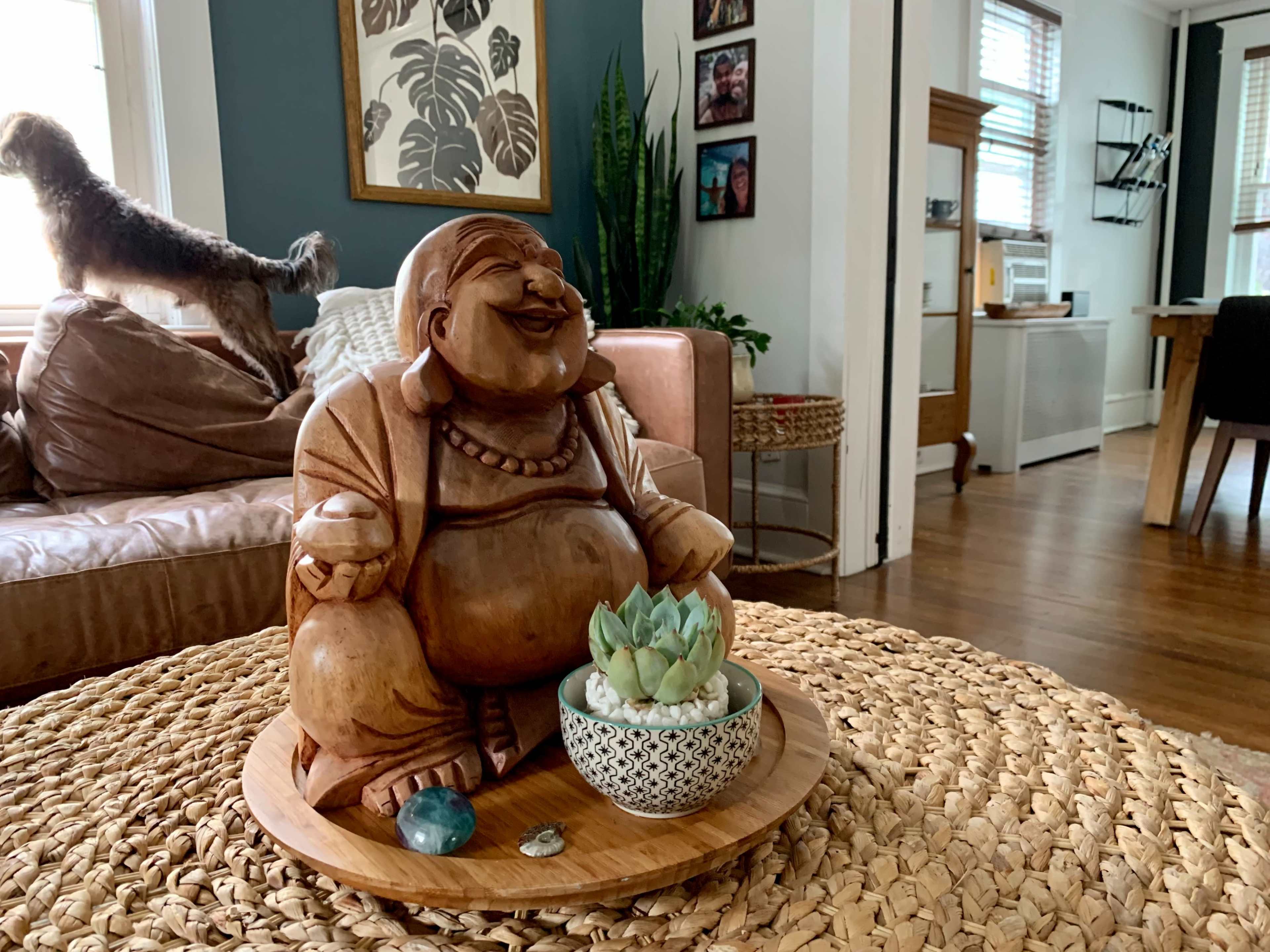 A wooden Buddha statue sits on a round tray with a succulent plant and gemstones, surrounded by a cozy living room.