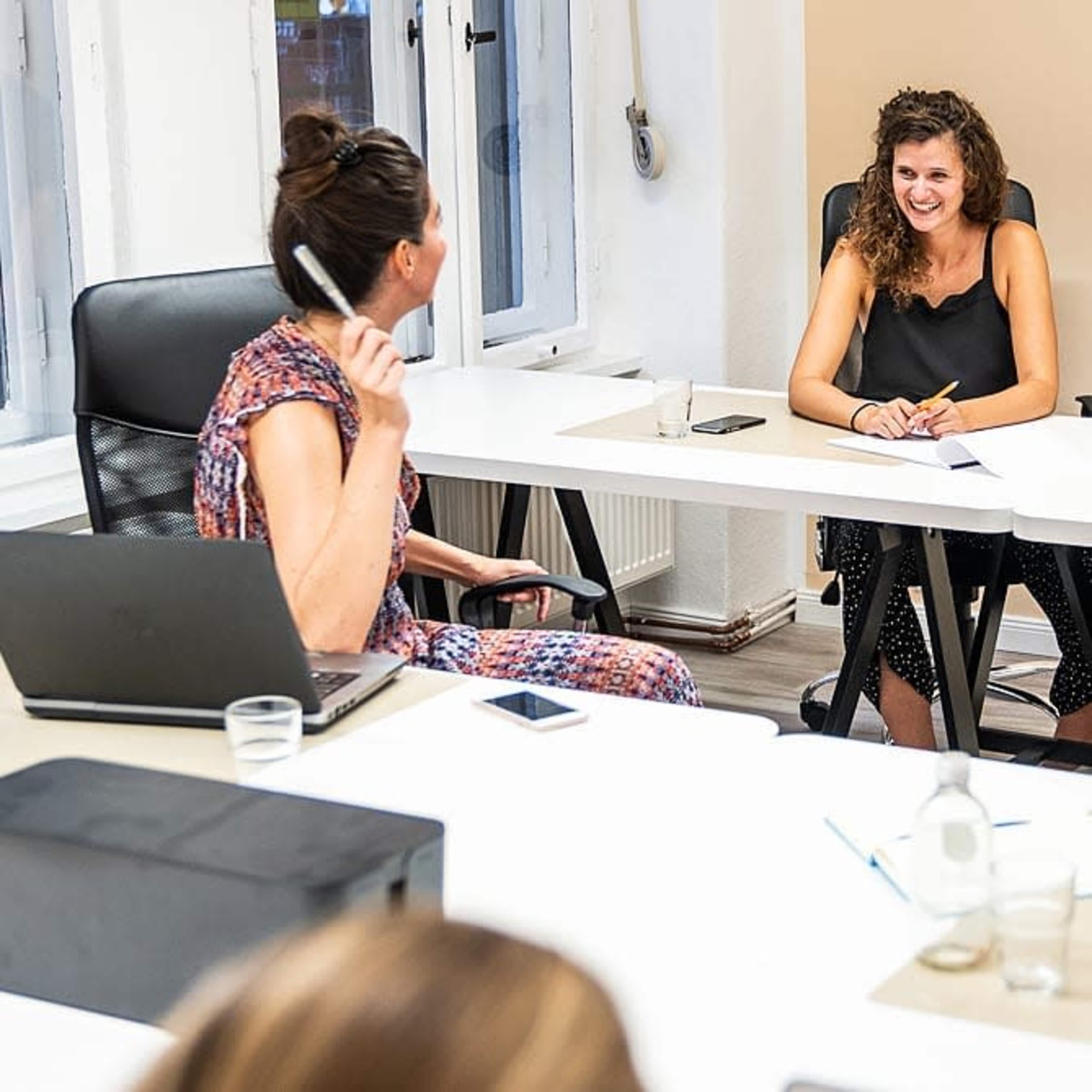 Two women are engaged in a discussion at a conference table in a brightly lit office, with laptops and notebooks visible.