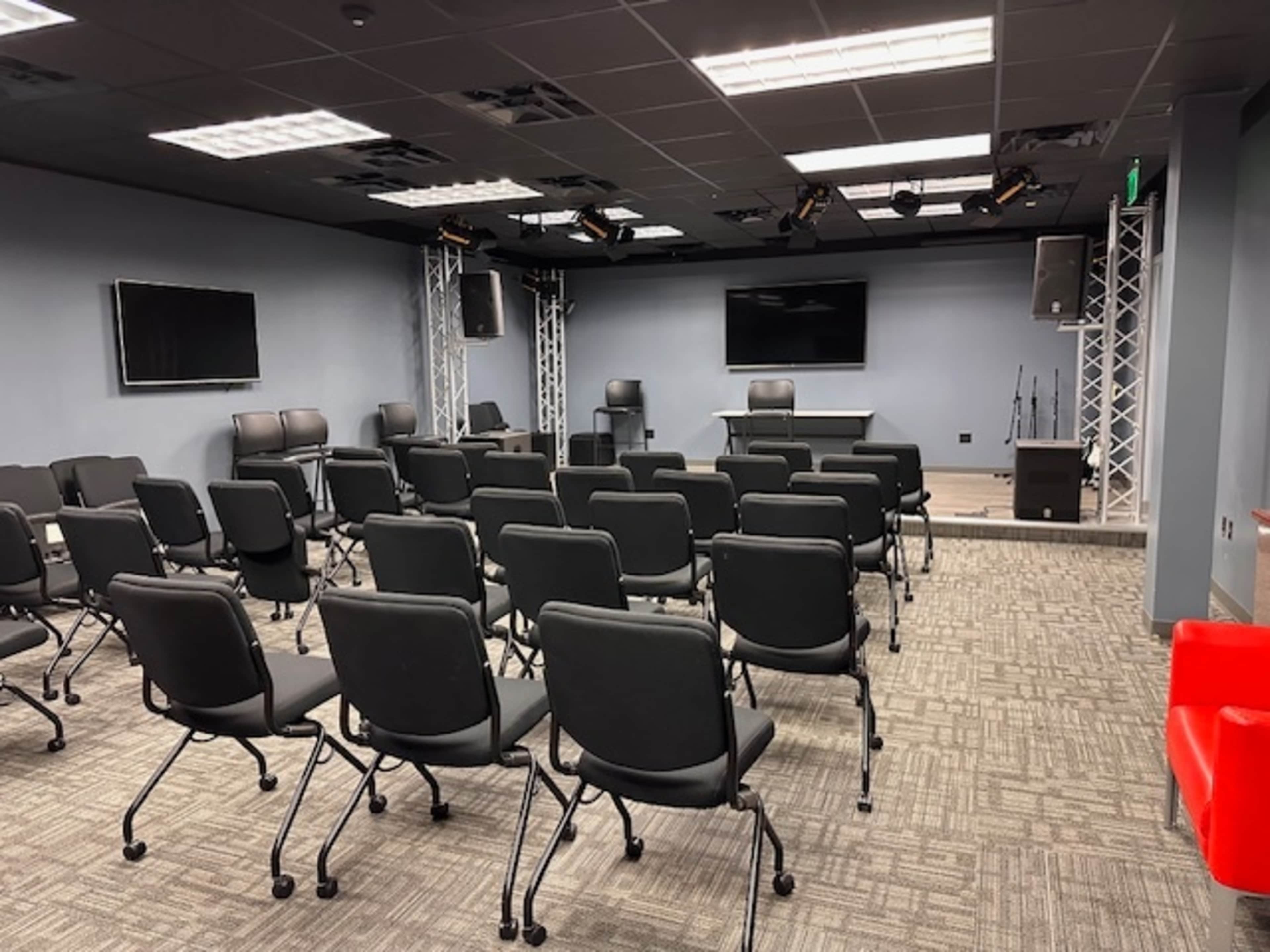 The image shows a modern conference room set up with rows of black chairs, a presentation area at the front, and audio-visual equipment.
