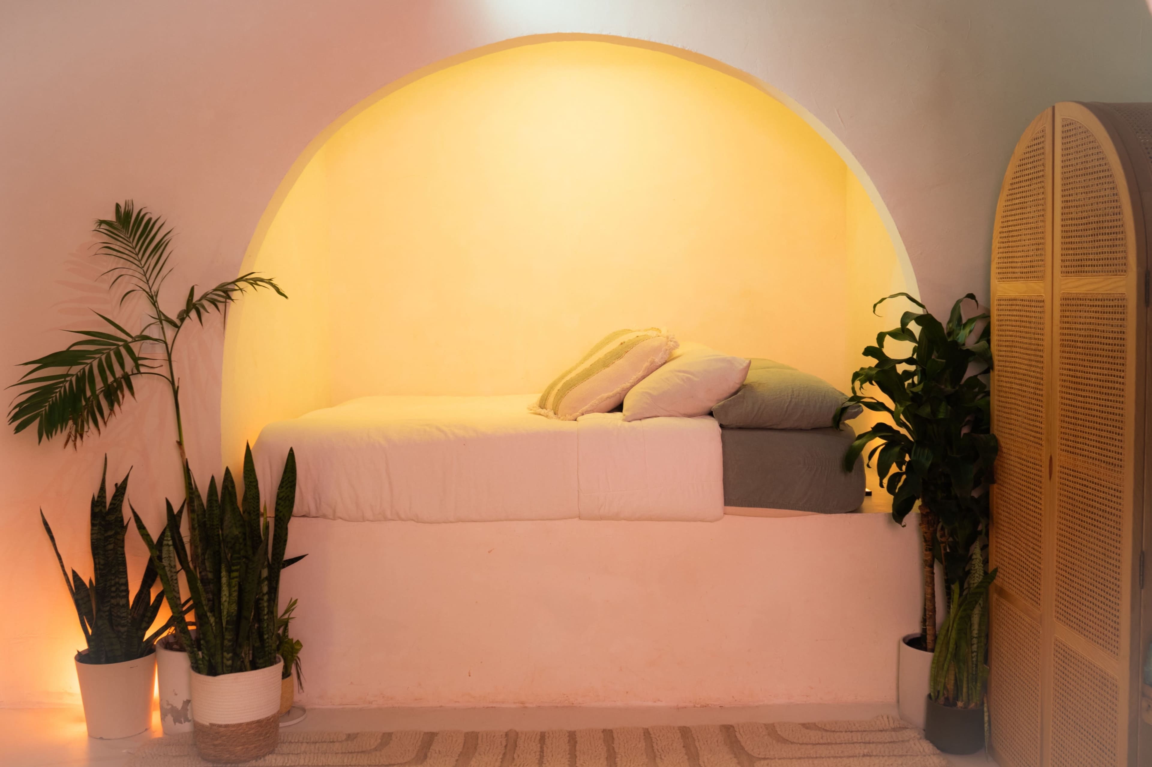A cozy alcove features a raised bed with soft pillows, surrounded by potted plants and illuminated by a warm light.