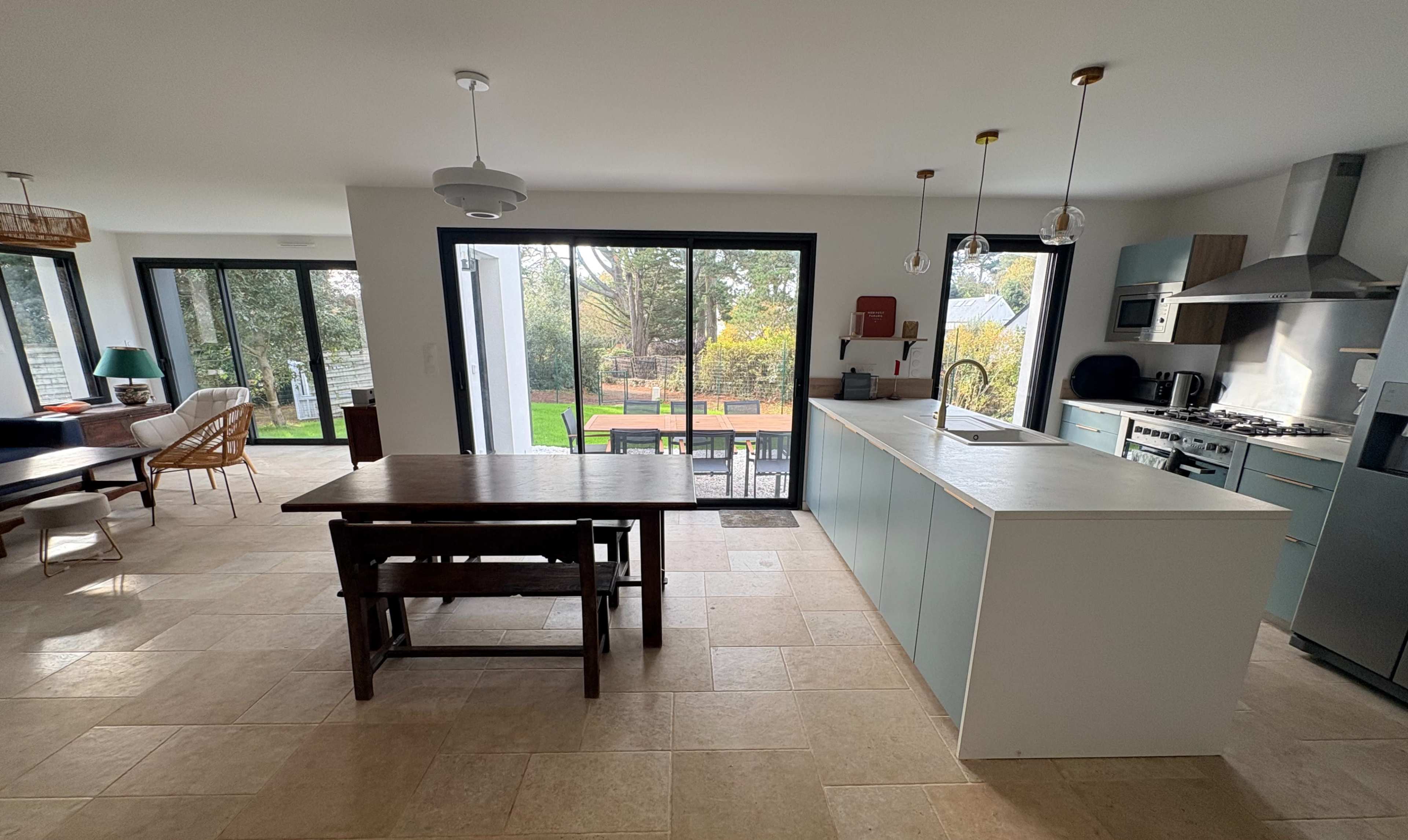 The image depicts a modern kitchen and dining area with a wooden table, large windows, and an open view of a garden outside.