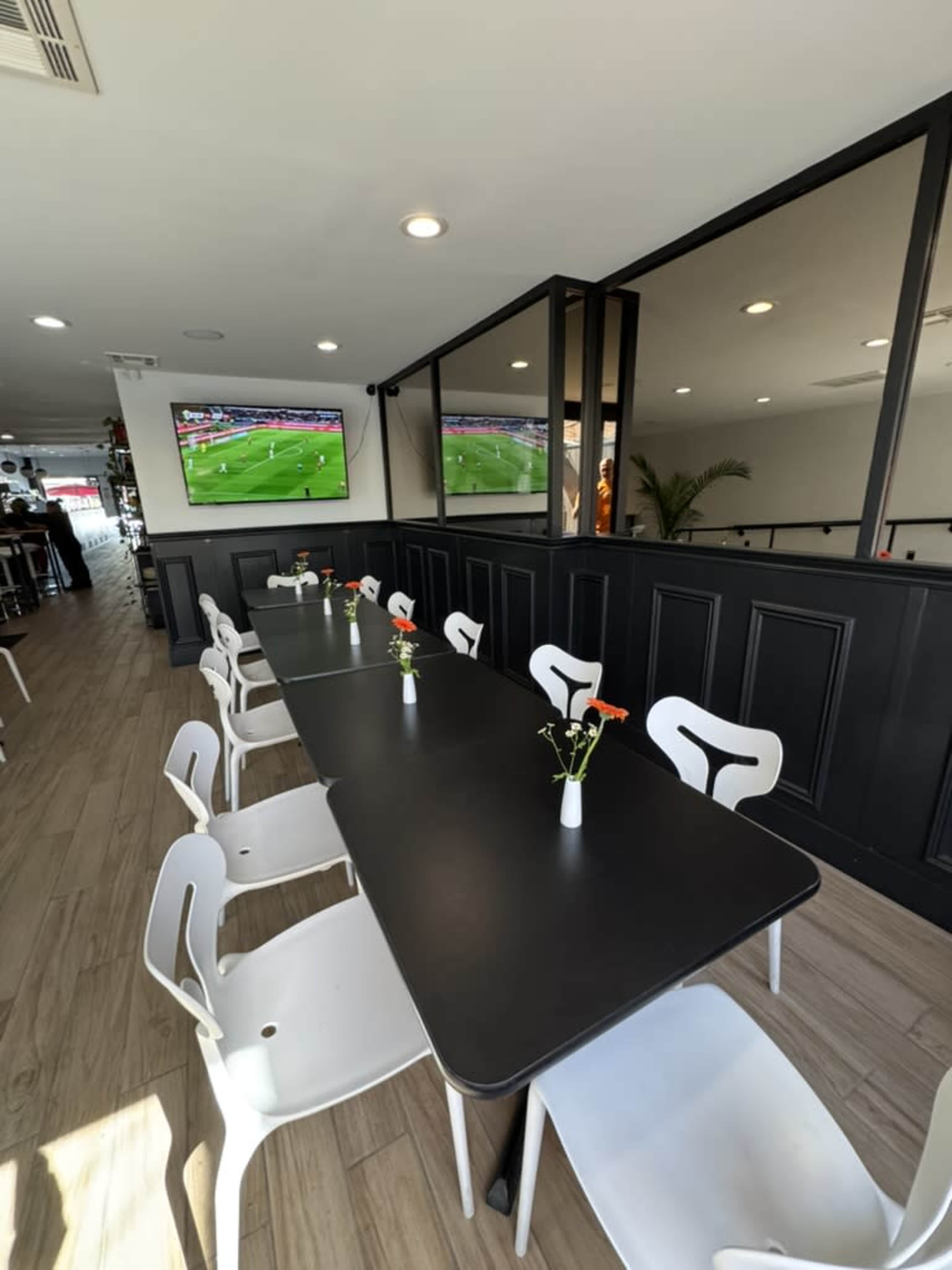 A modern restaurant interior features a long black table surrounded by white chairs, with small vases of flowers on the table and a large screen displaying a sports event in the background.