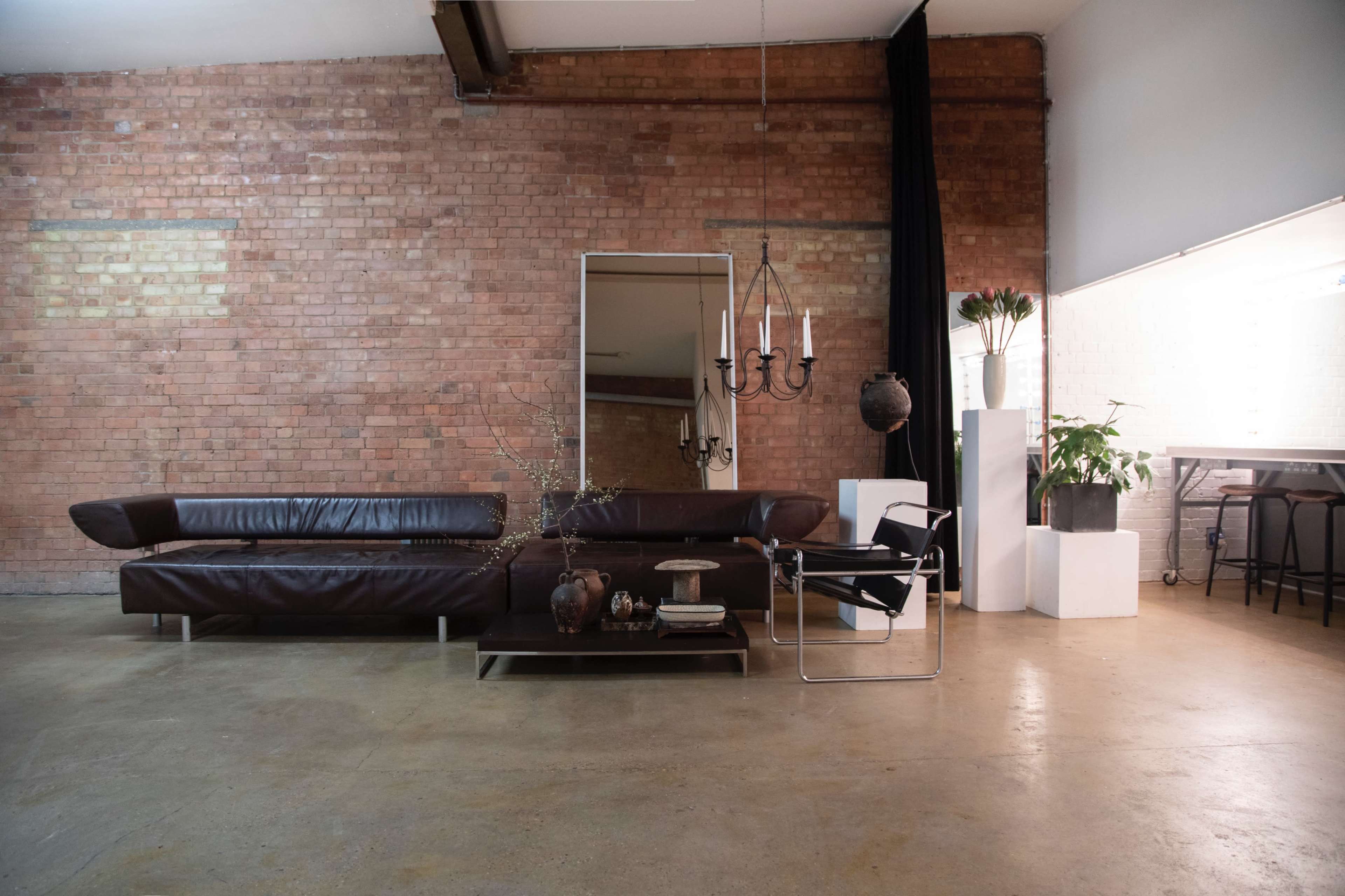 A modern living room featuring a leather sectional sofa, a glass coffee table, a contemporary chair, and a plant against a brick wall.