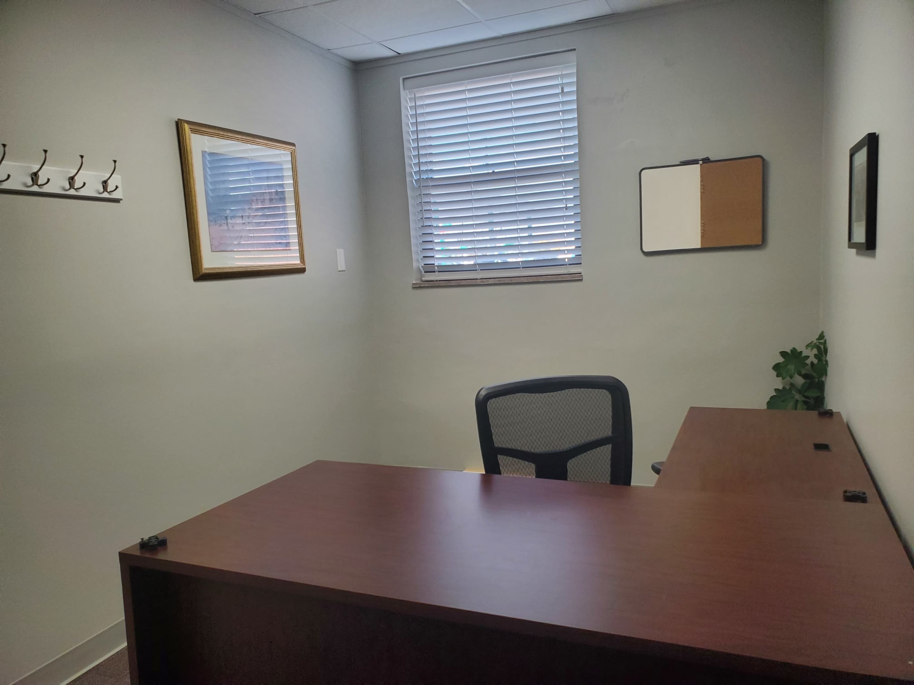 A small office with a wooden desk, a black chair, a window with blinds, and some wall-mounted coat hooks.