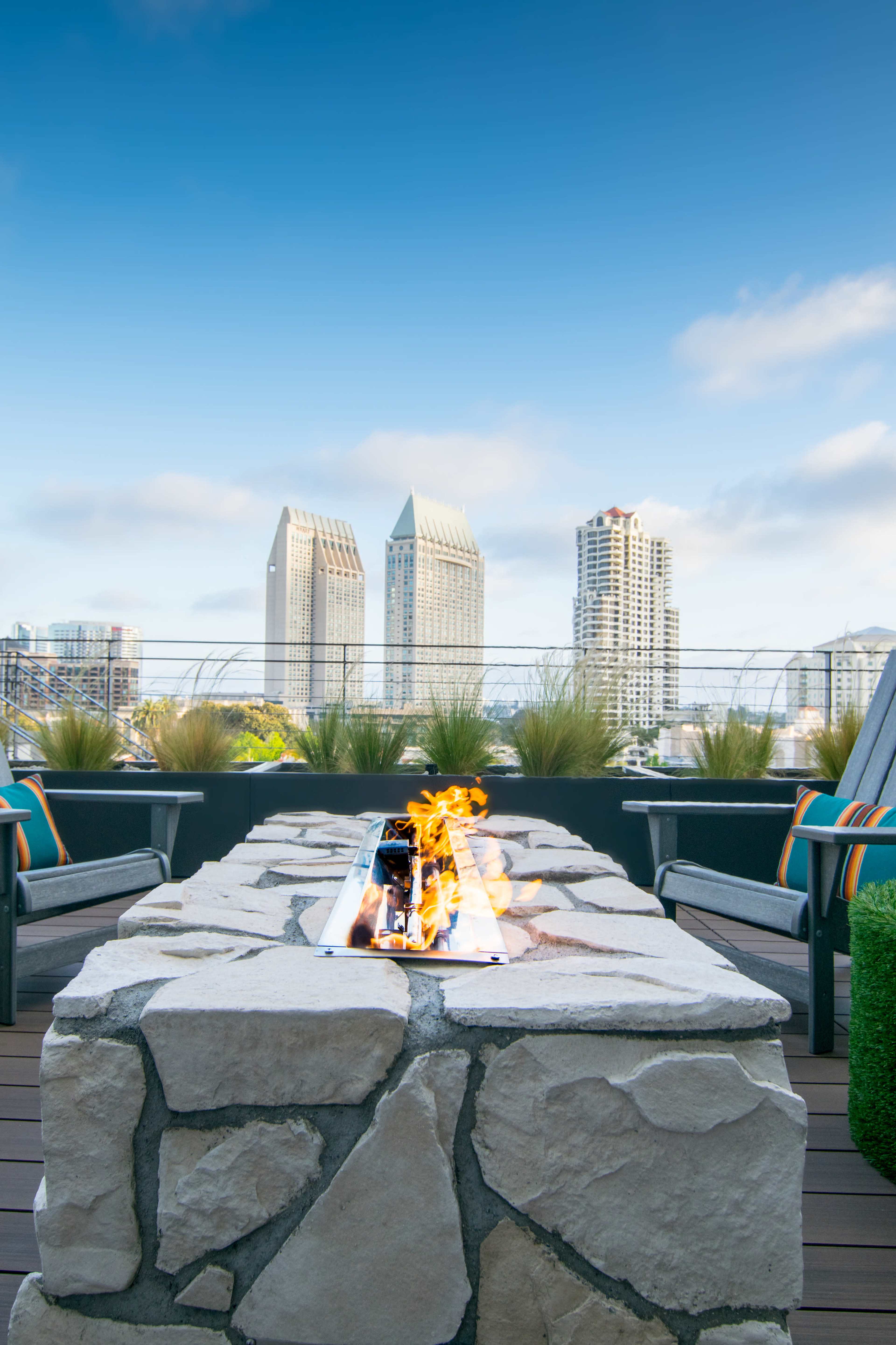 A rooftop patio features a stone fire pit with flames, surrounded by chairs and offering a view of tall buildings against a clear blue sky.