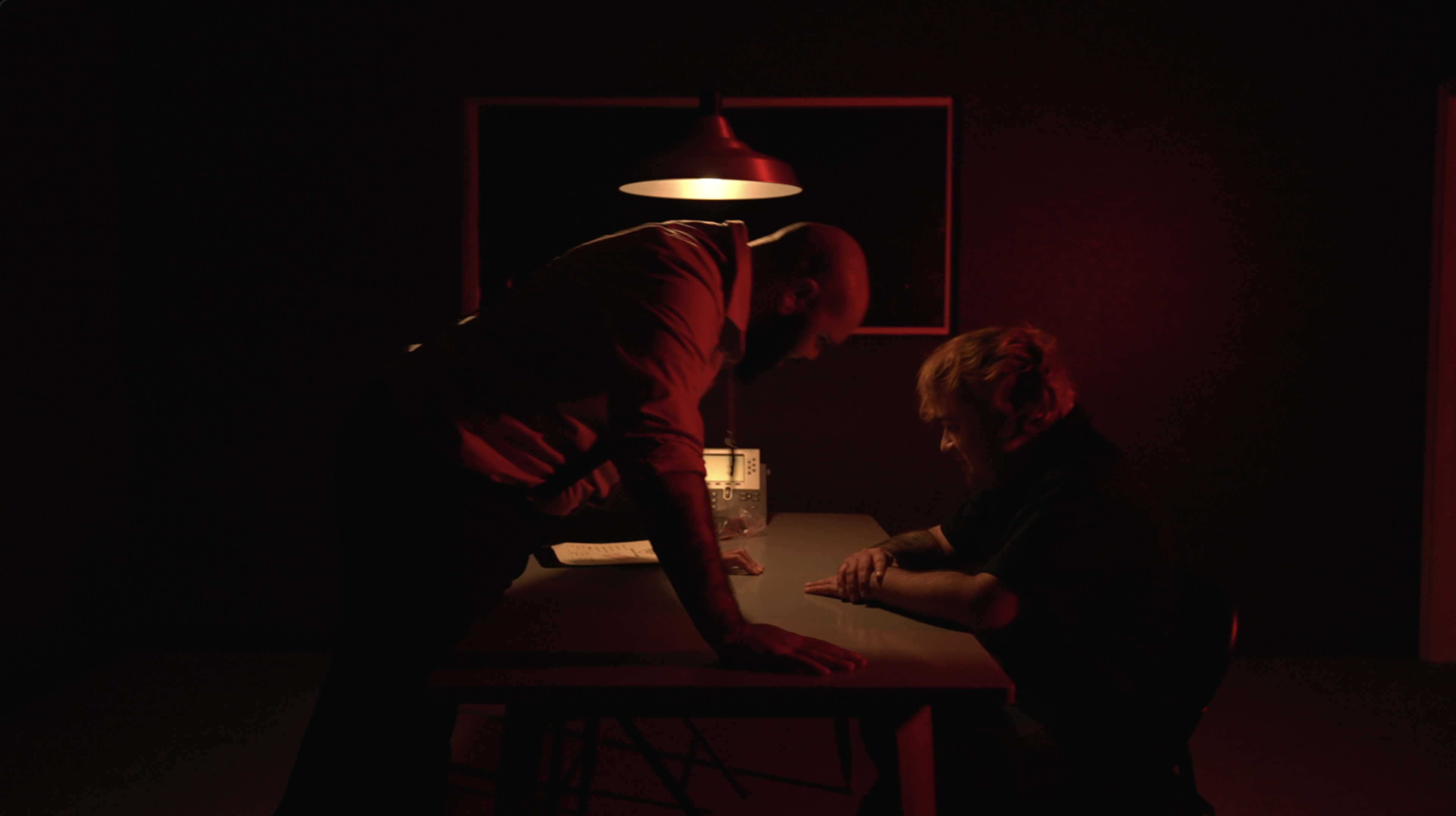 A tense interrogation scene is set in a dimly lit room with a man leaning over a table towards another seated figure.