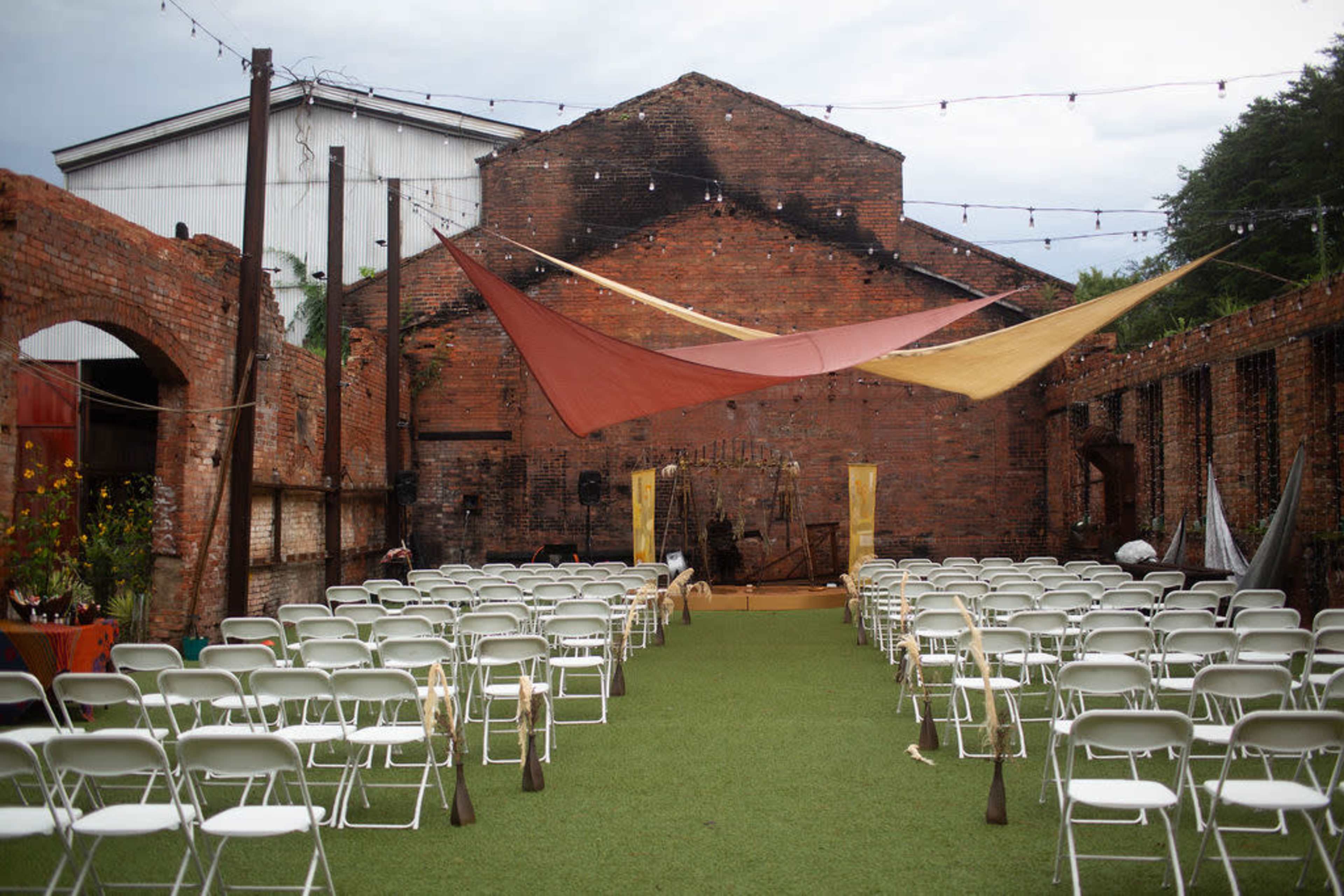 An outdoor venue features rows of white chairs arranged on green grass facing a small stage, framed by rustic brick walls and draped fabric overhead.