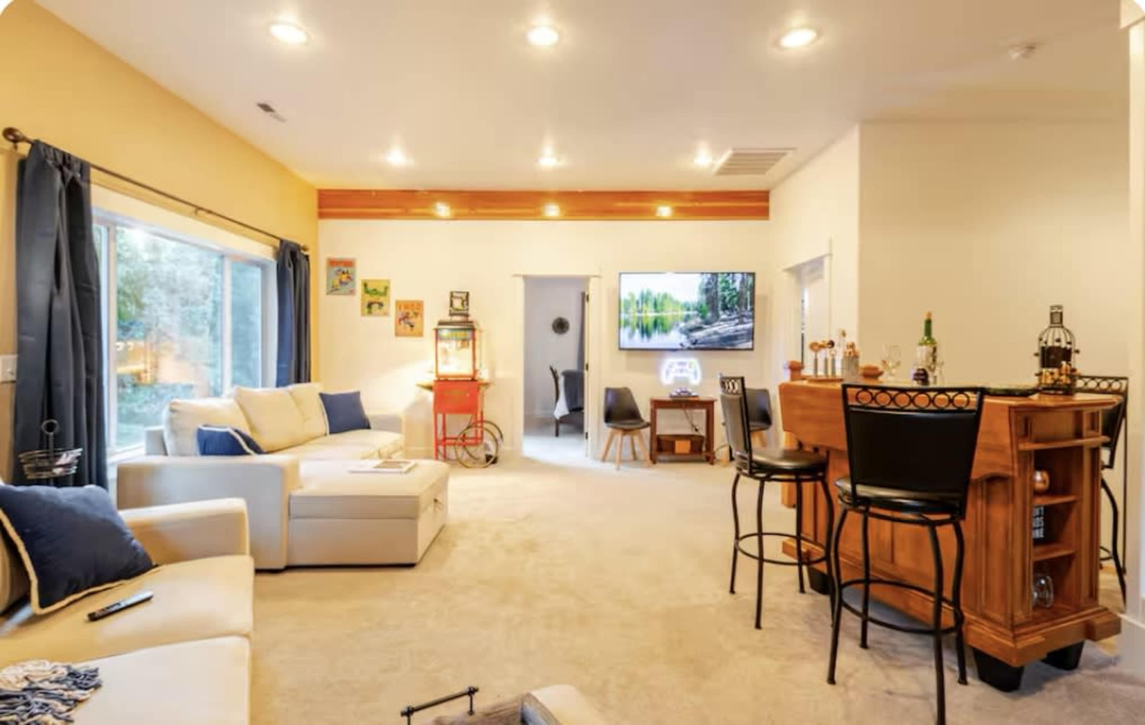 The image shows a spacious living room with a bar area, a comfortable sofa, and a large screen television mounted on the wall.