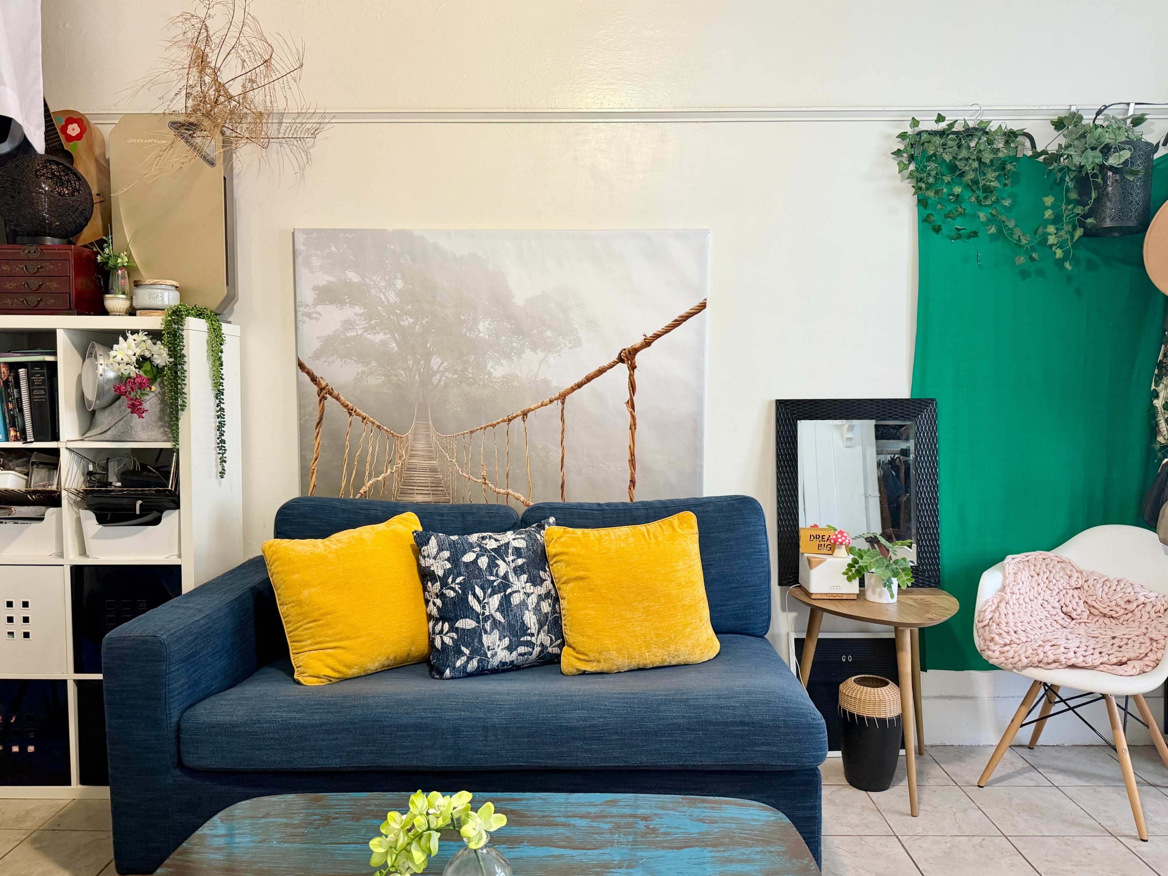 A blue sofa with colorful pillows is positioned against a wall featuring a large scenic print, surrounded by plants and decorative items in a cozy living space.