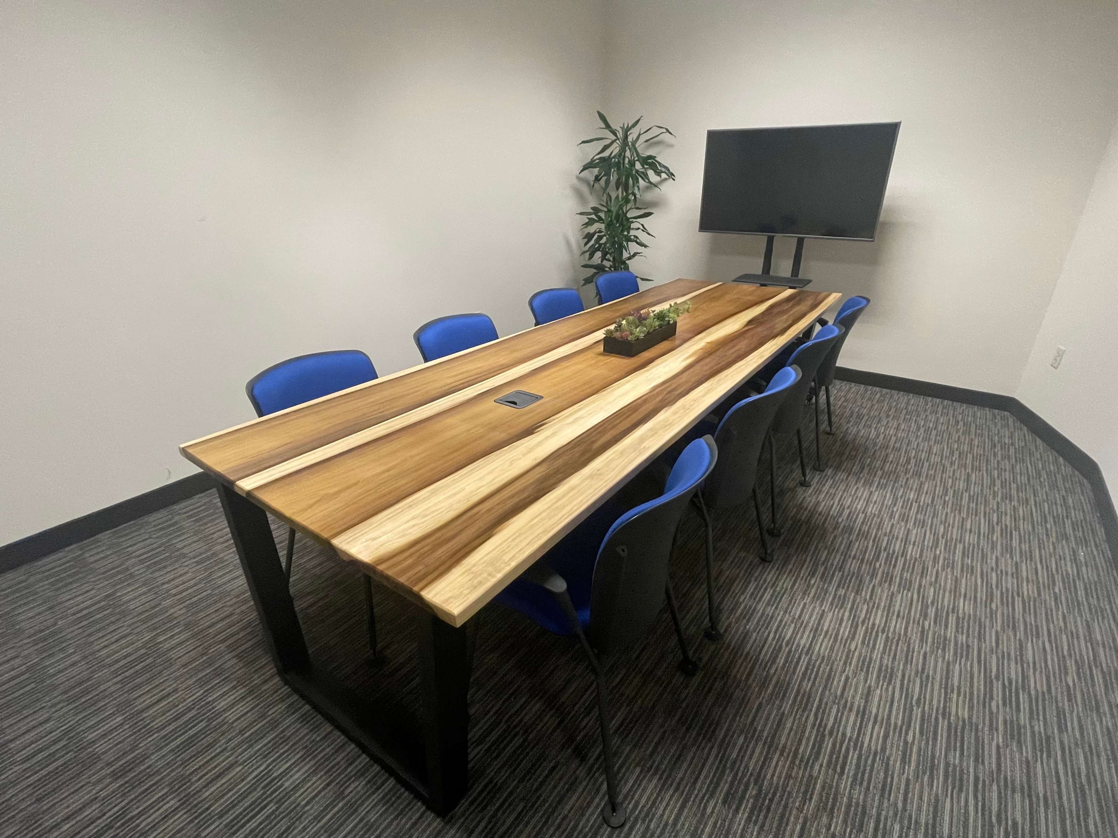 A conference room features a large wooden table surrounded by eight blue chairs and equipped with a television mounted on the wall.