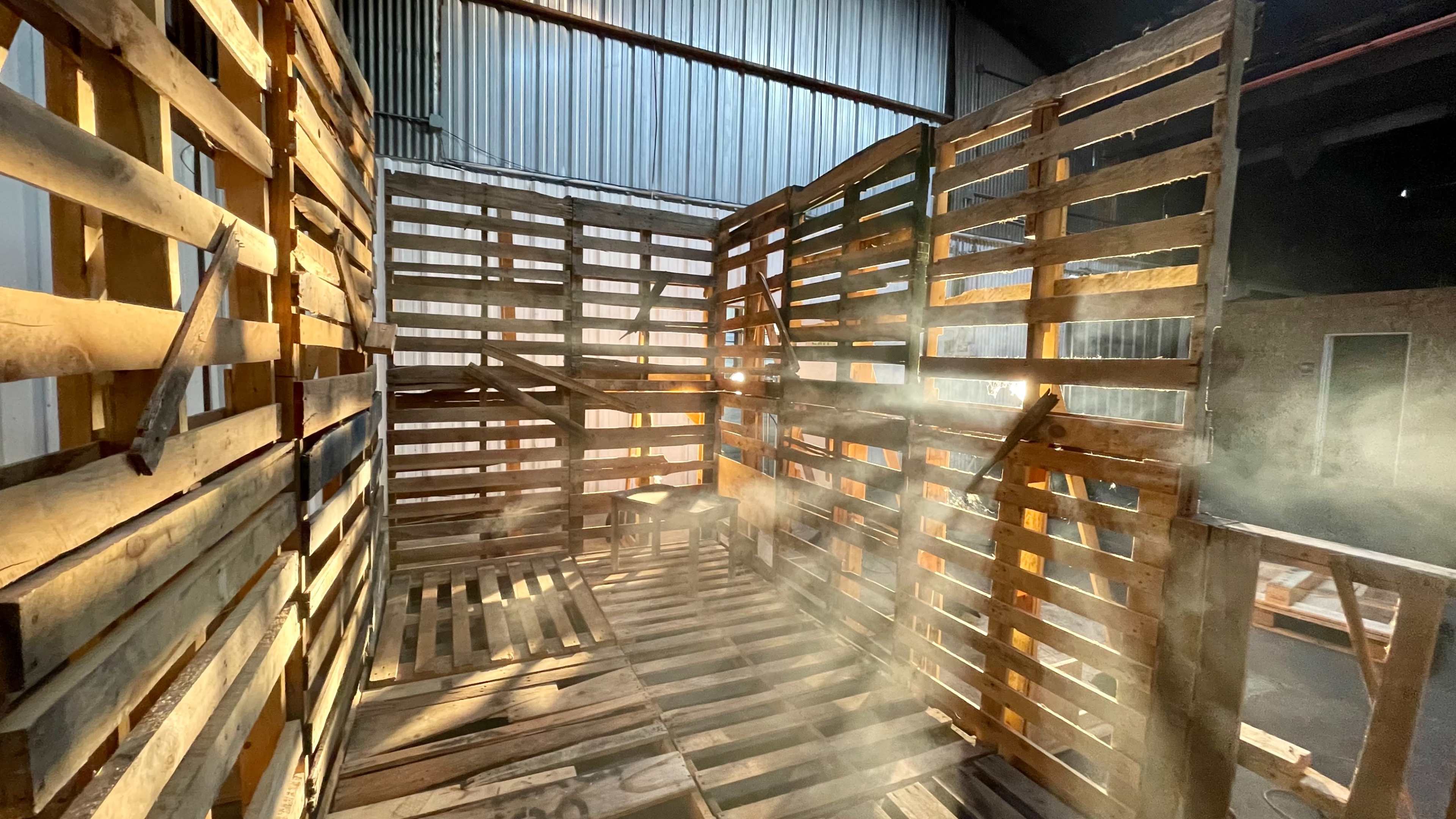 An interior space constructed from wooden pallets, illuminated by streams of light filtering through and creating a smoky atmosphere.