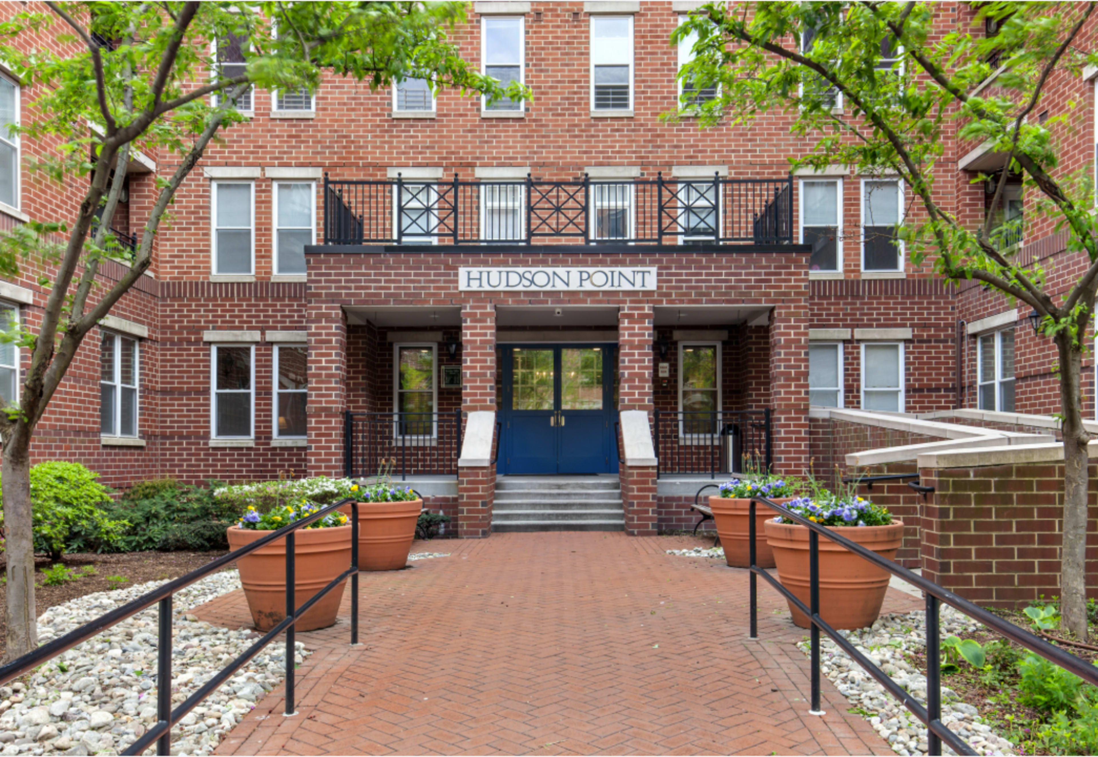The entrance to Hudson Point features a brick facade with potted plants on either side of a walkway leading to a blue door.