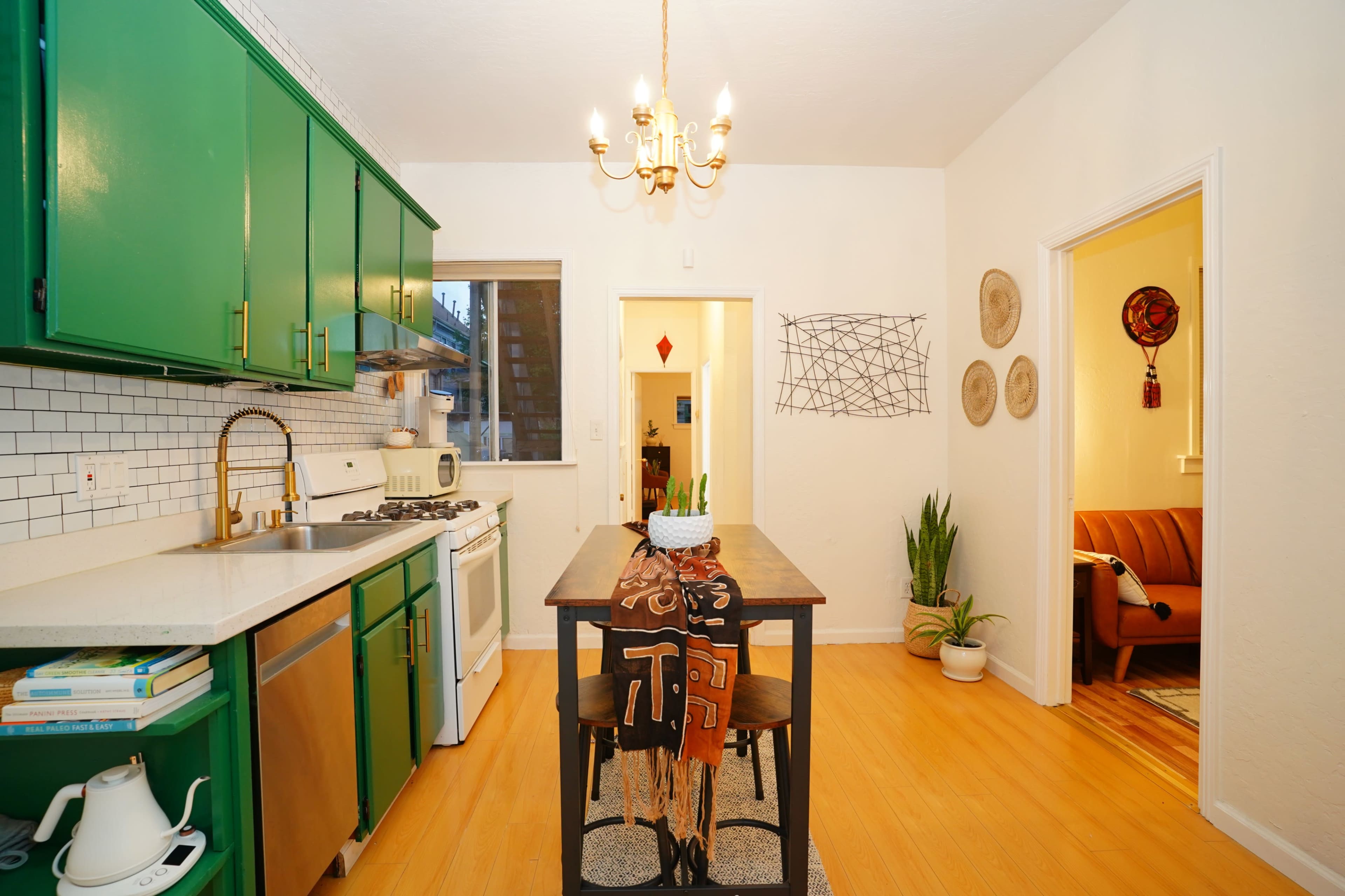 The image shows a kitchen with green cabinets, a white countertop, a central table with two chairs, and an adjoining living area visible through an open doorway.