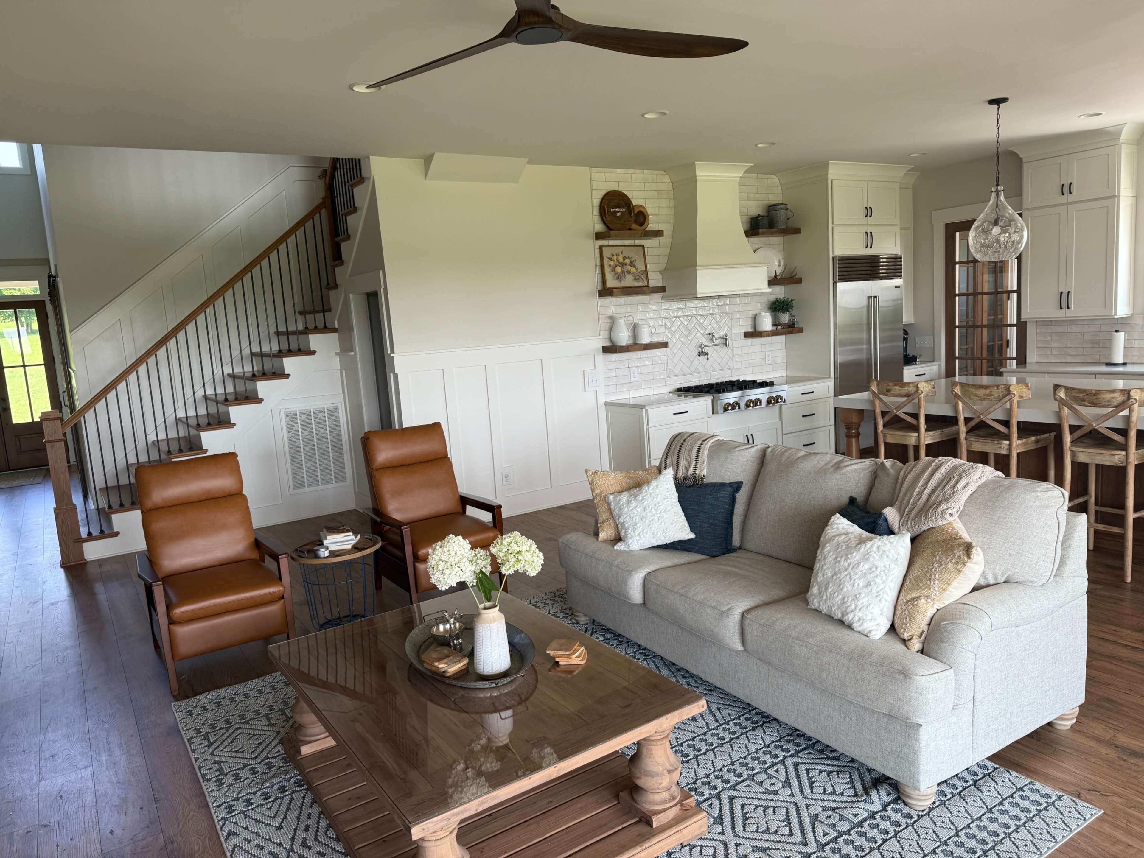 The image shows a modern living room with a light-colored sofa, two brown armchairs, a wooden coffee table, and a staircase leading to a second level, all set in an open floor plan adjacent to a kitchen.