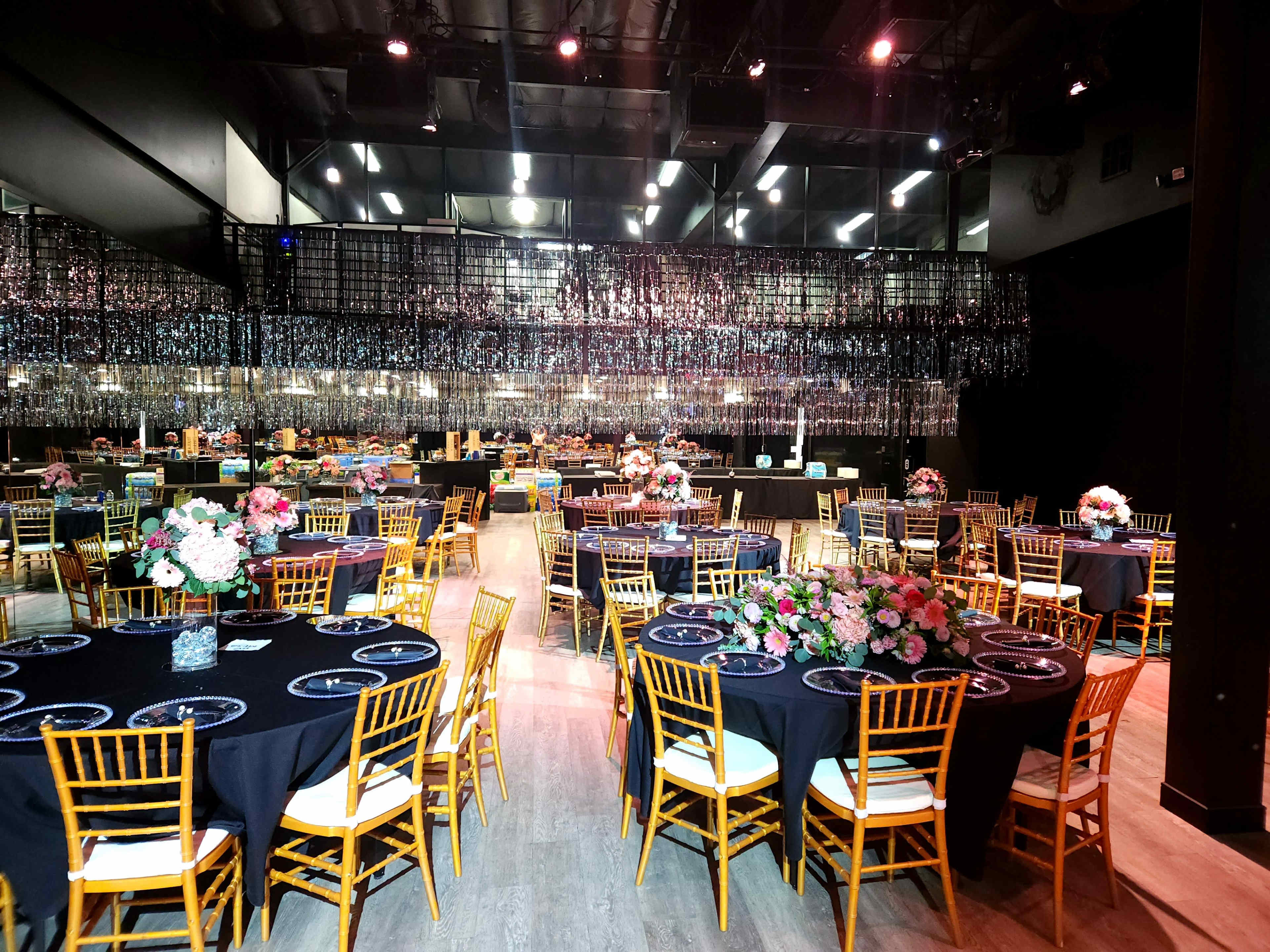 The image shows a spacious banquet hall set for an event, with round tables covered in black cloth and decorated with floral centerpieces.