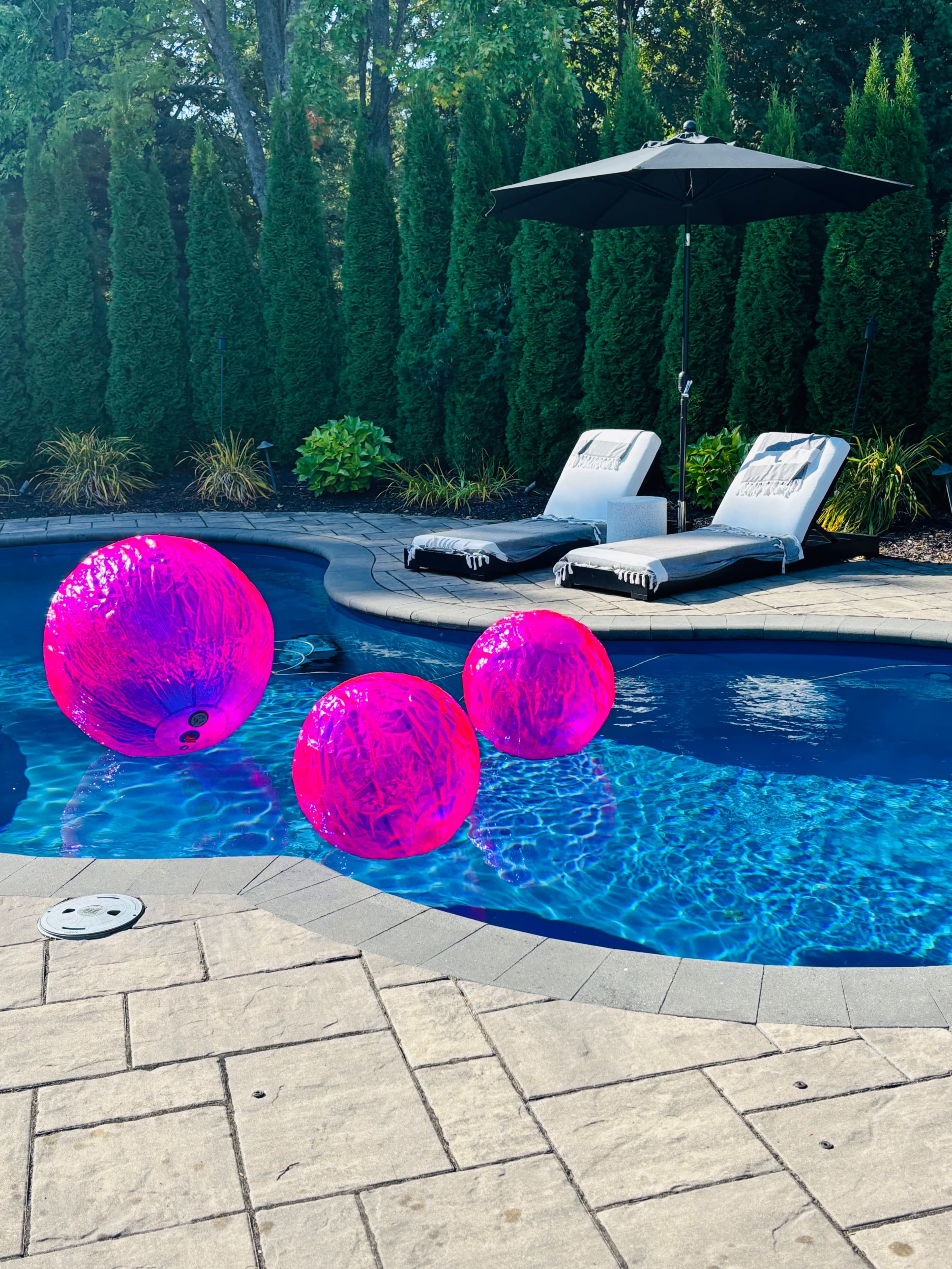 The image shows a swimming pool surrounded by tall green hedges, featuring three large pink balls floating on the water and two lounge chairs beside the pool under an umbrella.