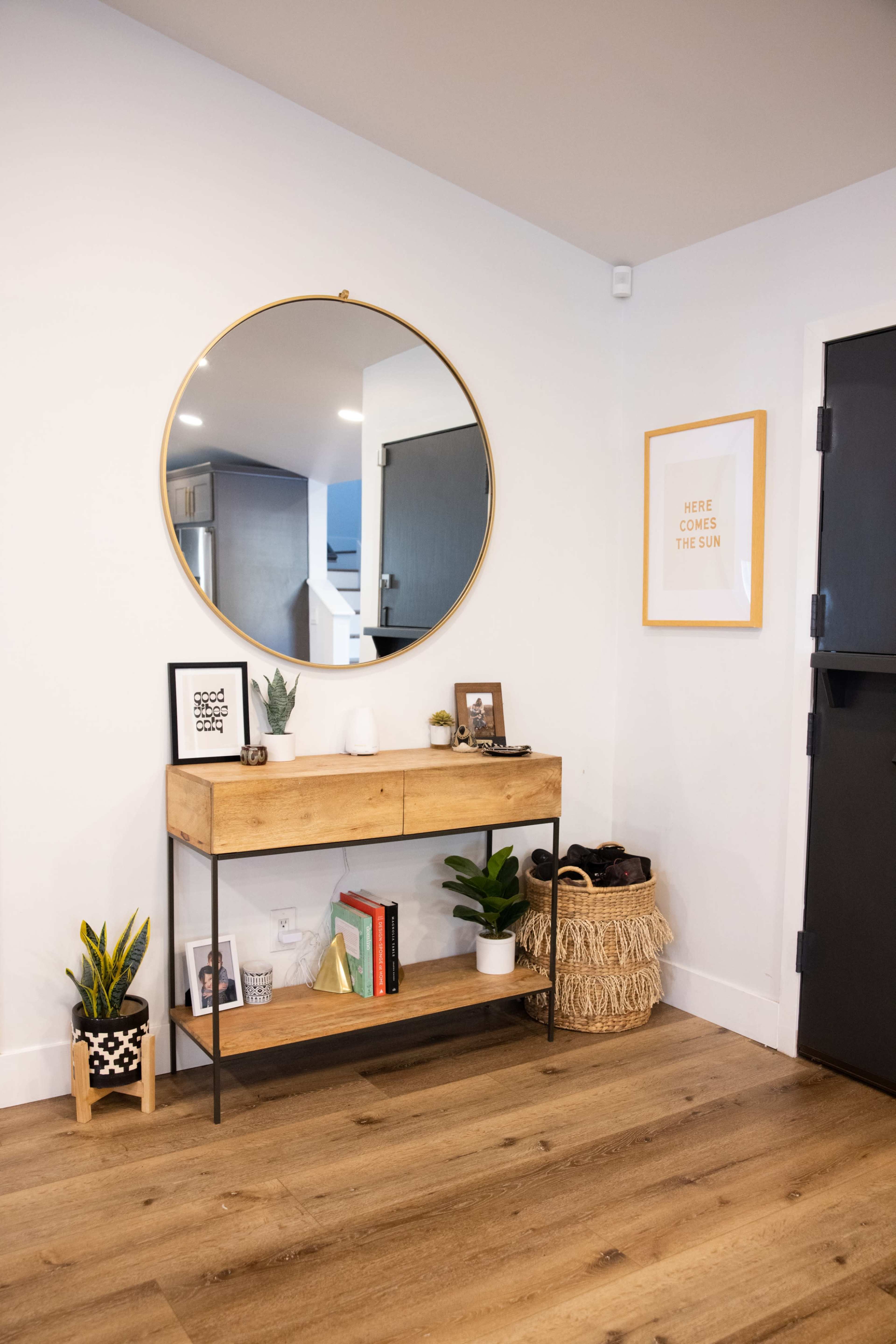 A wooden console table with a round mirror above it is positioned against a white wall, featuring decorative plants and various items on the shelves.