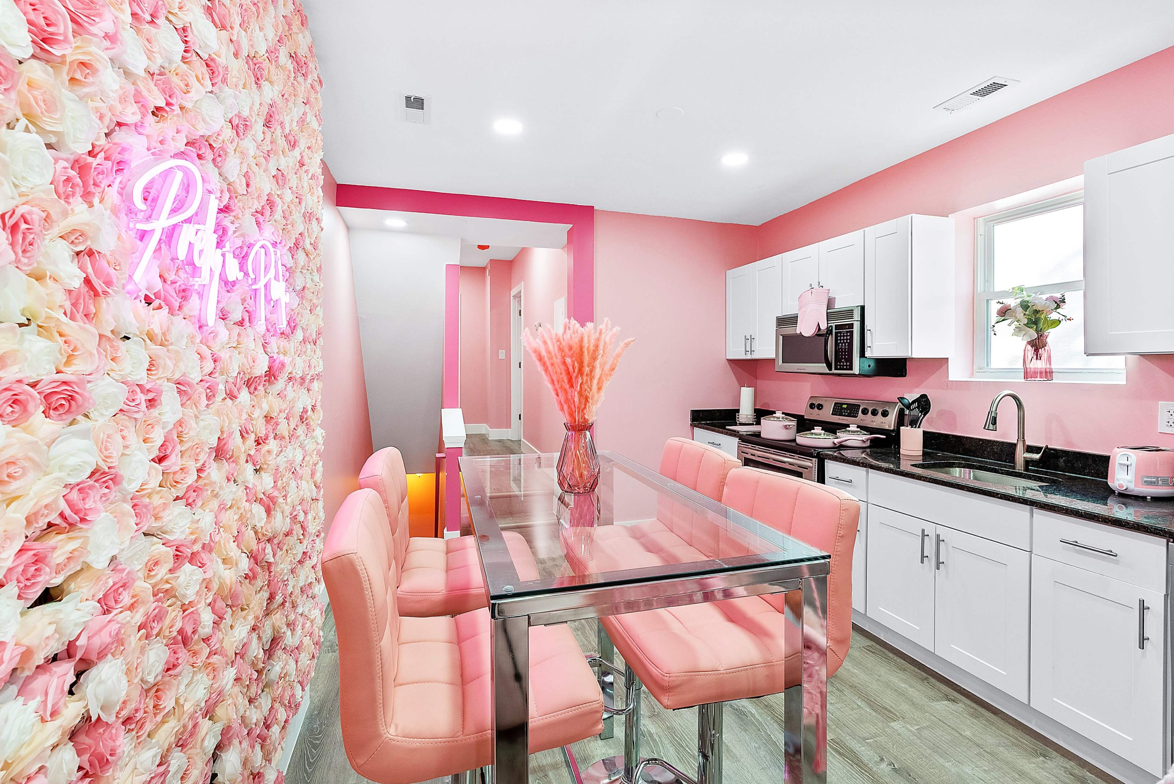 A bright kitchen with a pink floral wall, a glass dining table surrounded by pink chairs, and modern white cabinetry.