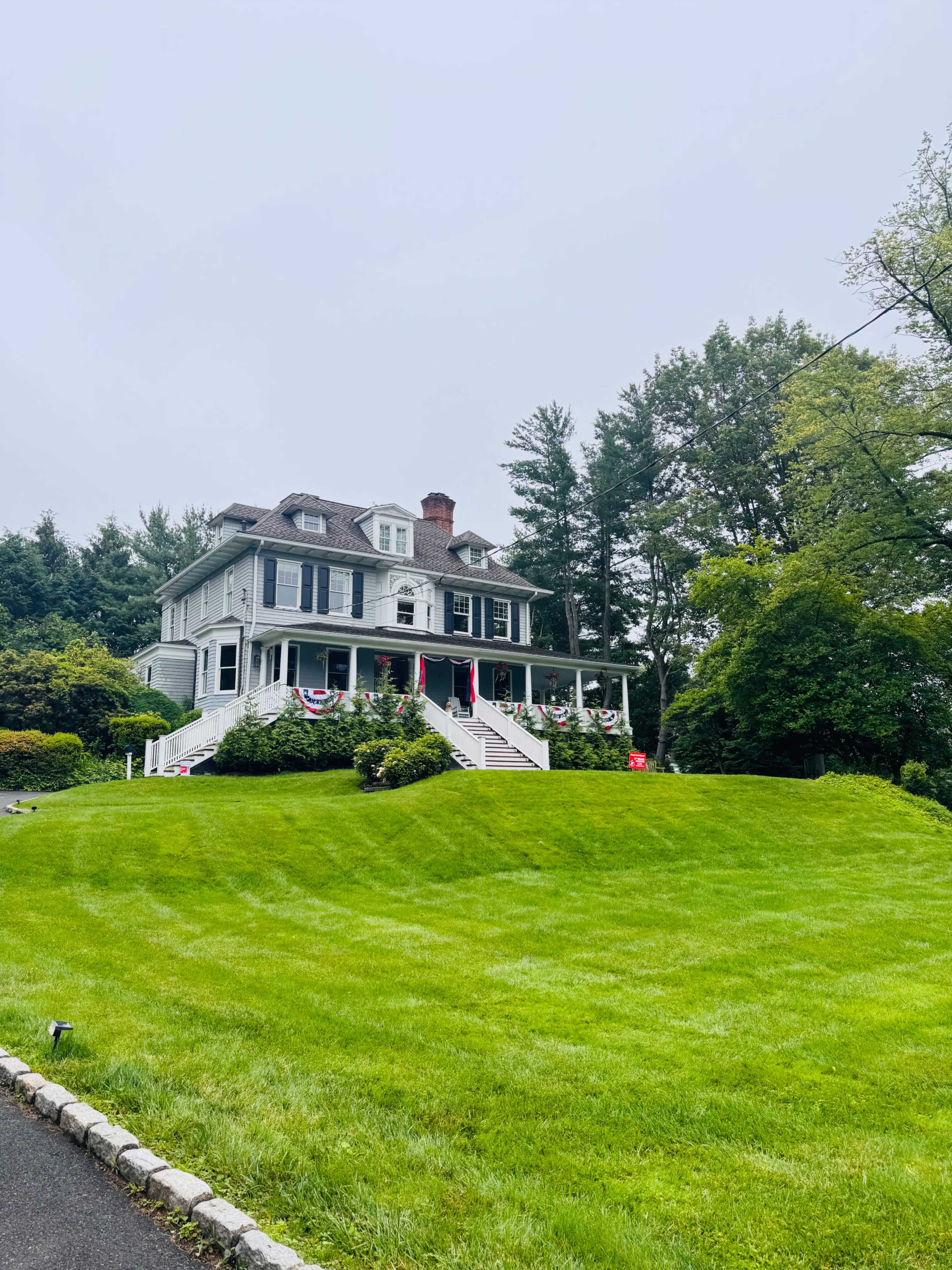 A large, two-story house with a gray exterior and white trim sits on a grassy hill, surrounded by trees, and features decorative red, white, and blue bunting on its porch.