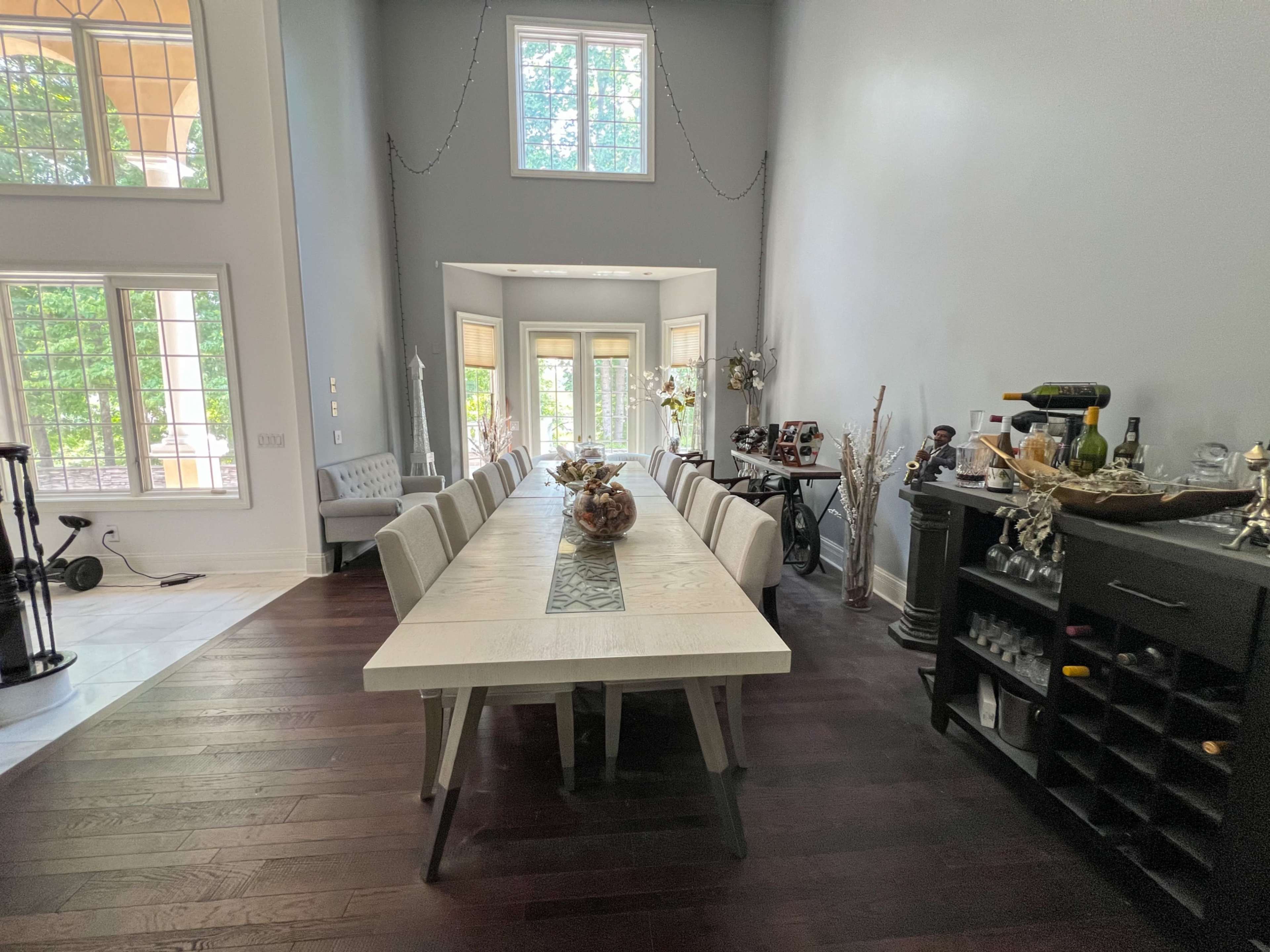 The image shows a spacious dining area with a long table and chairs arranged along a hardwood floor, flanked by large windows and a bar cart in the corner.