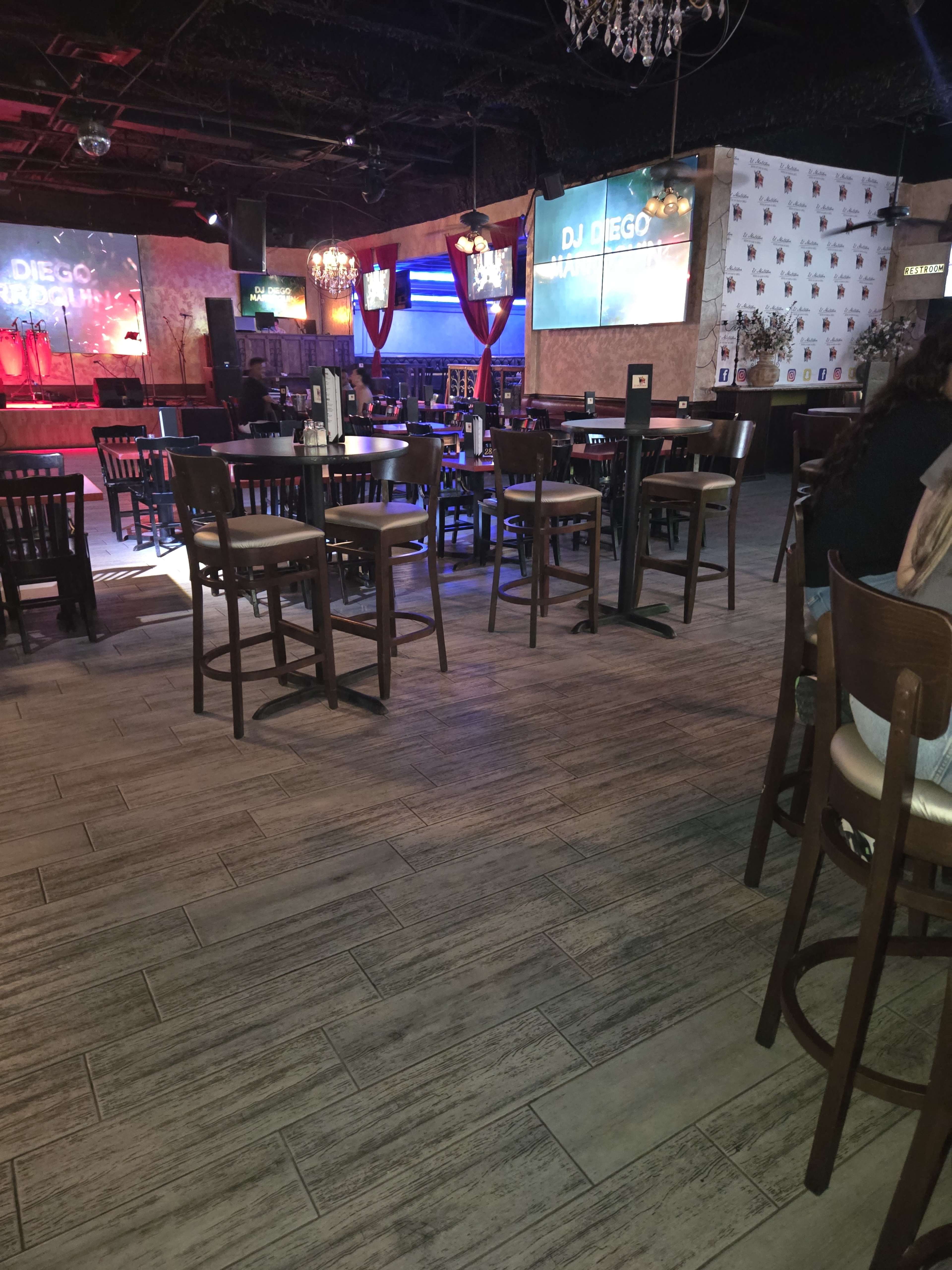 The image shows a partially empty bar or restaurant venue with tables and chairs arranged around a stage area, where a screen displays "DJ DIEGO."