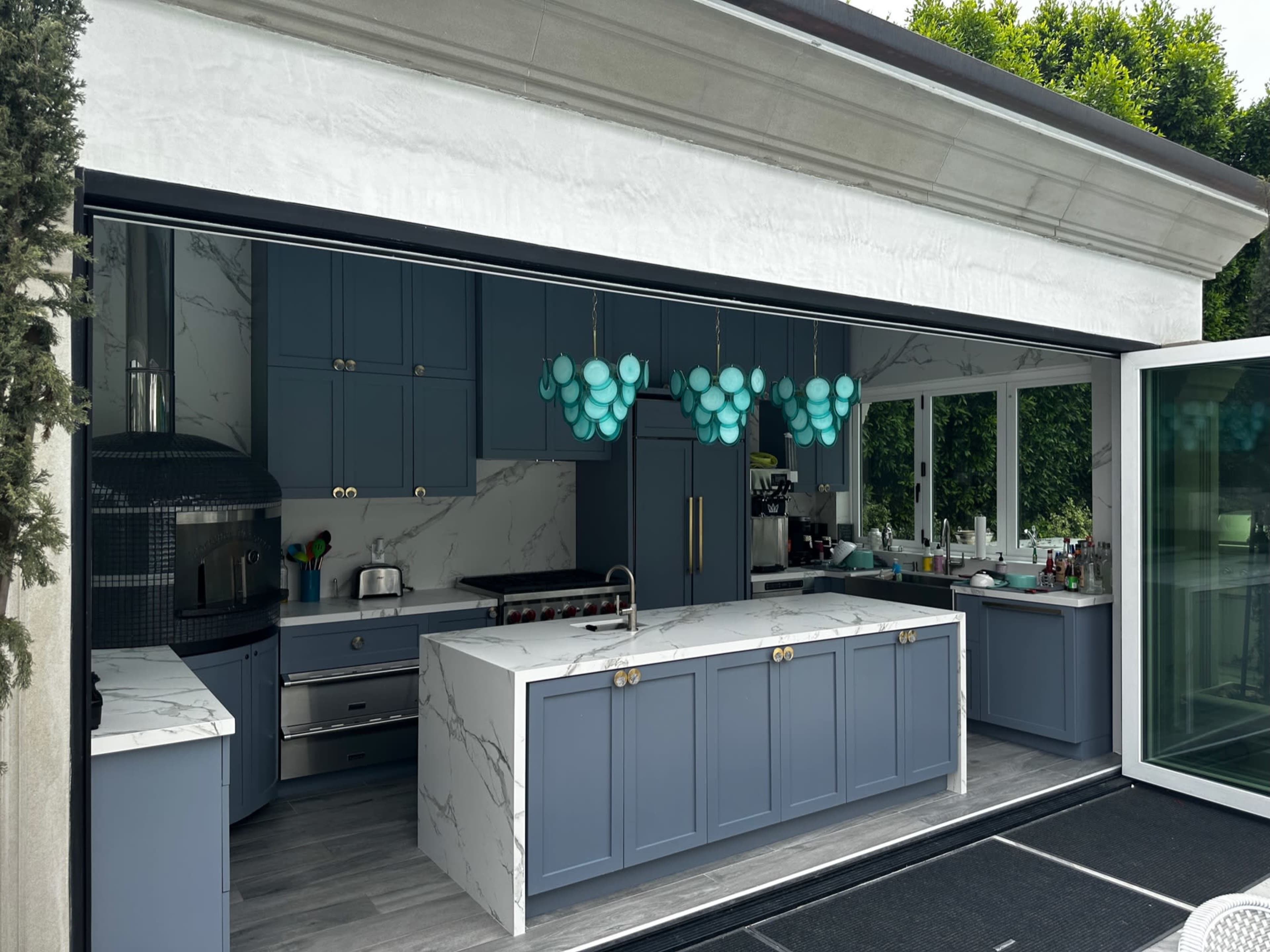 A modern kitchen with blue cabinetry, a large marble island, and turquoise pendant lights, integrated into an open layout with sliding glass doors.