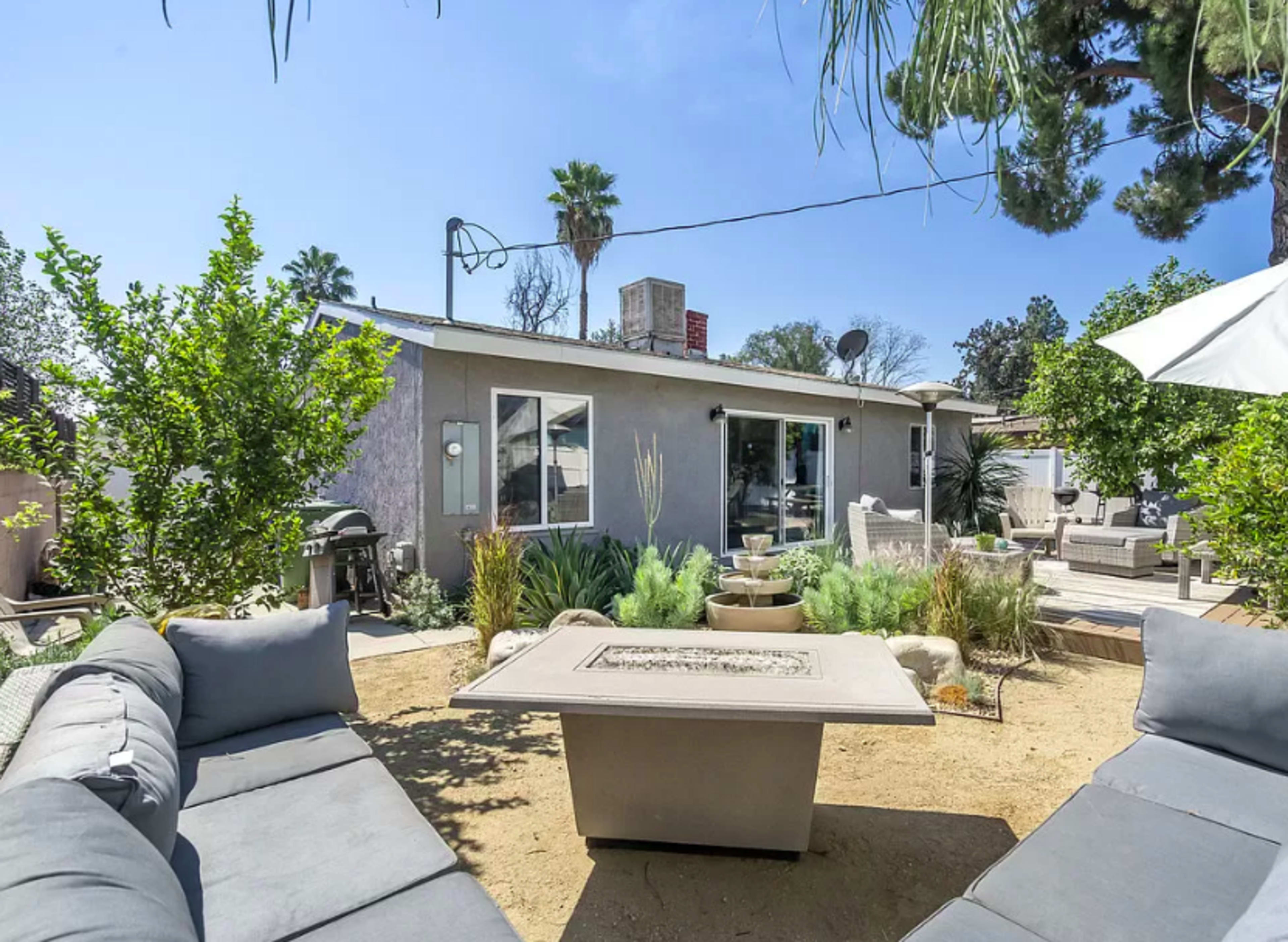 A backyard with a modern seating area surrounding a stone table, featuring various plants and a house in the background.