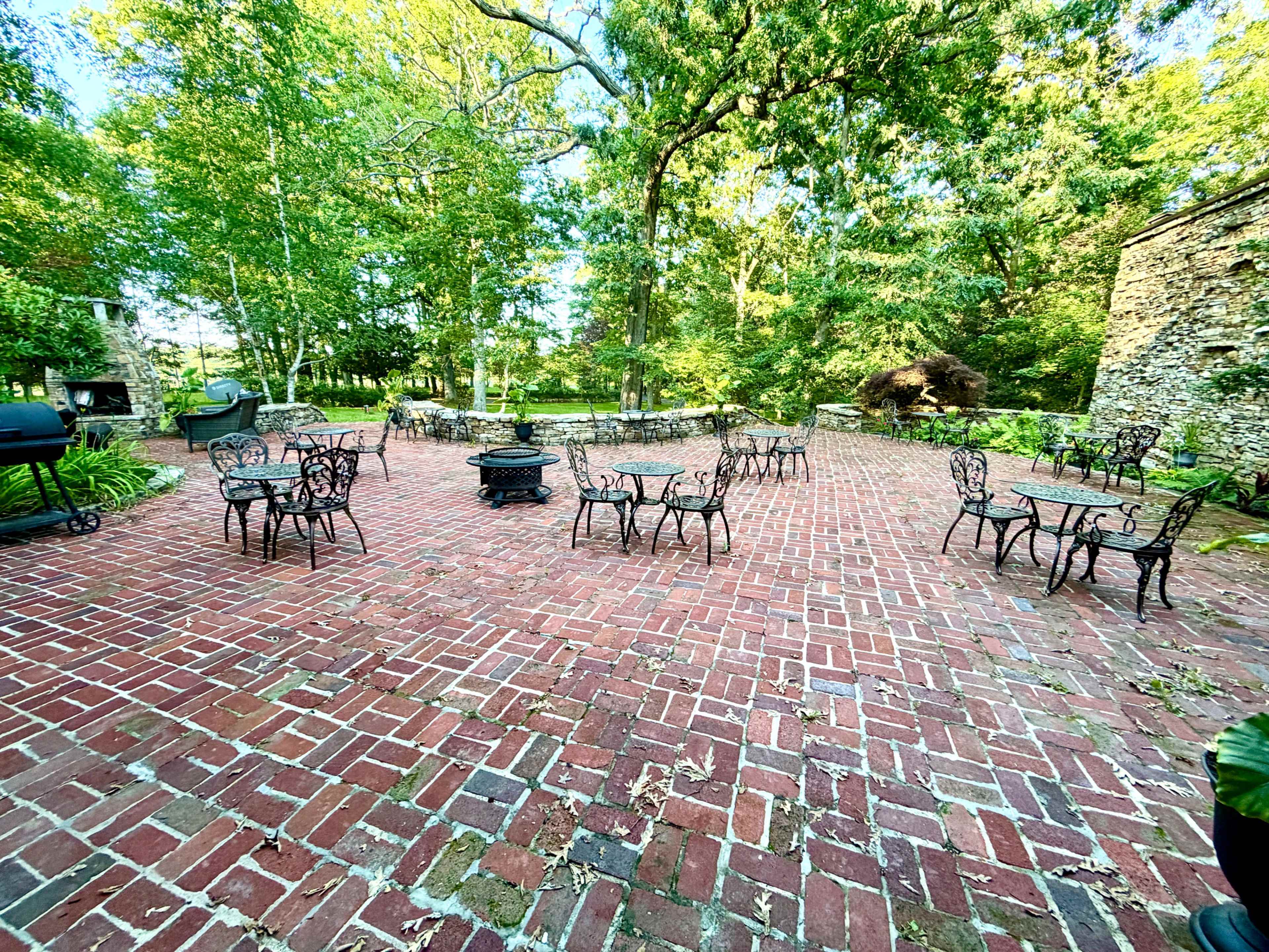 The image shows a brick patio surrounded by trees, featuring several metal tables and chairs arranged in an outdoor setting.
