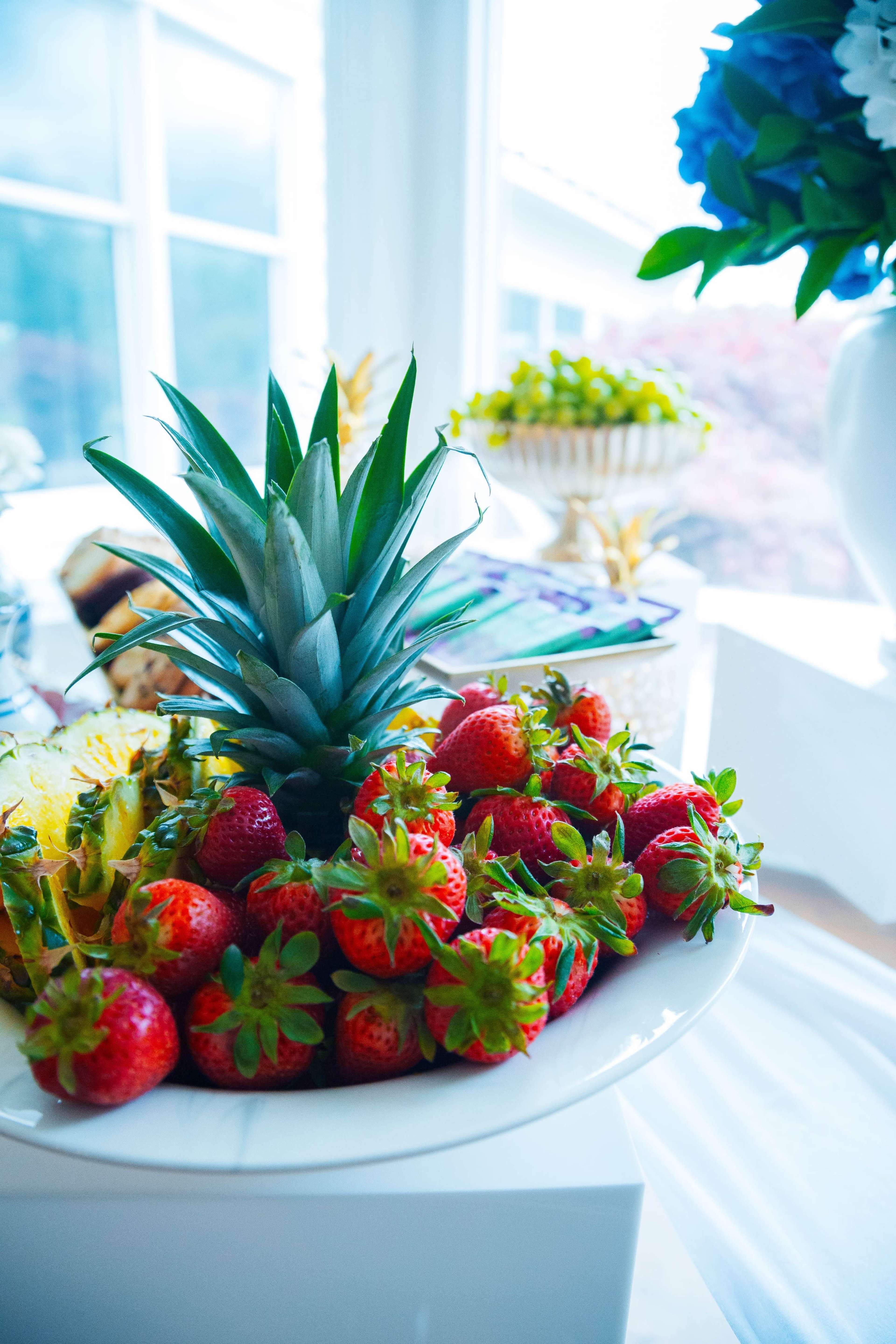 A plate filled with fresh strawberries and topped with a pineapple sits on a table by a window.