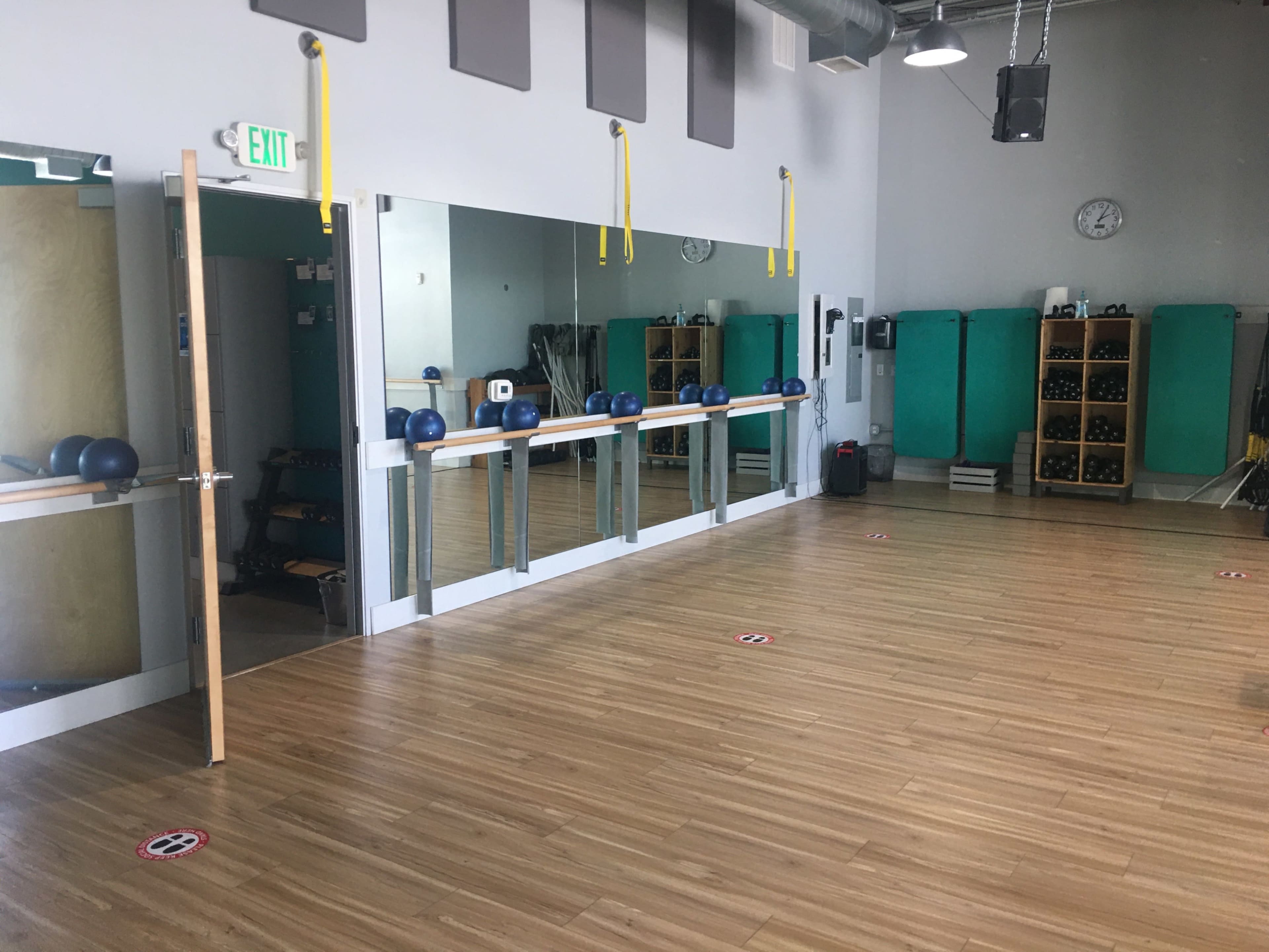 A fitness studio with wooden flooring, large mirrors, exercise equipment on shelves, and social distancing markers on the floor.