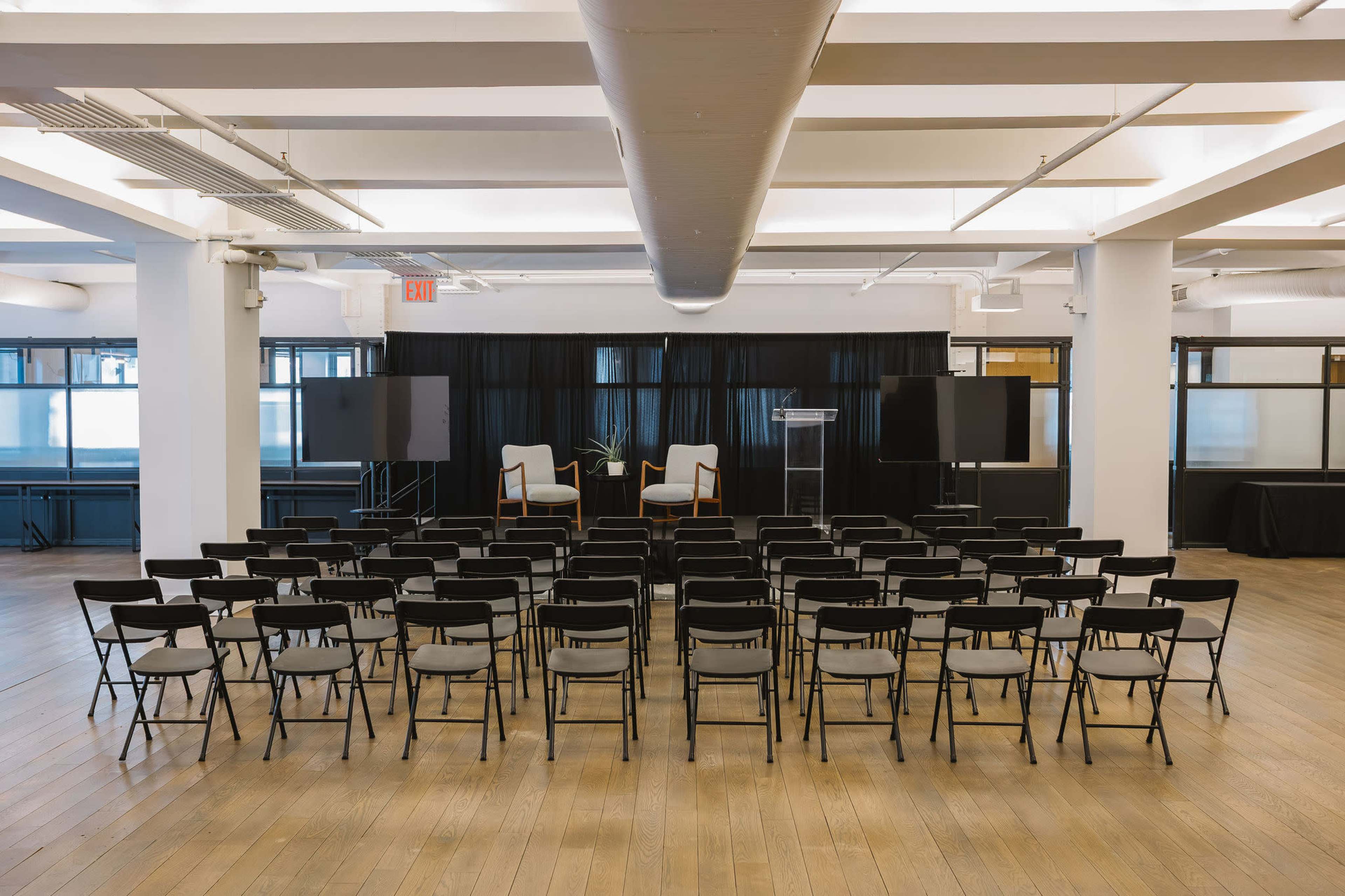 The image shows a spacious room set up for an event, featuring rows of black folding chairs facing a small stage with two chairs and a podium, flanked by blank screens.