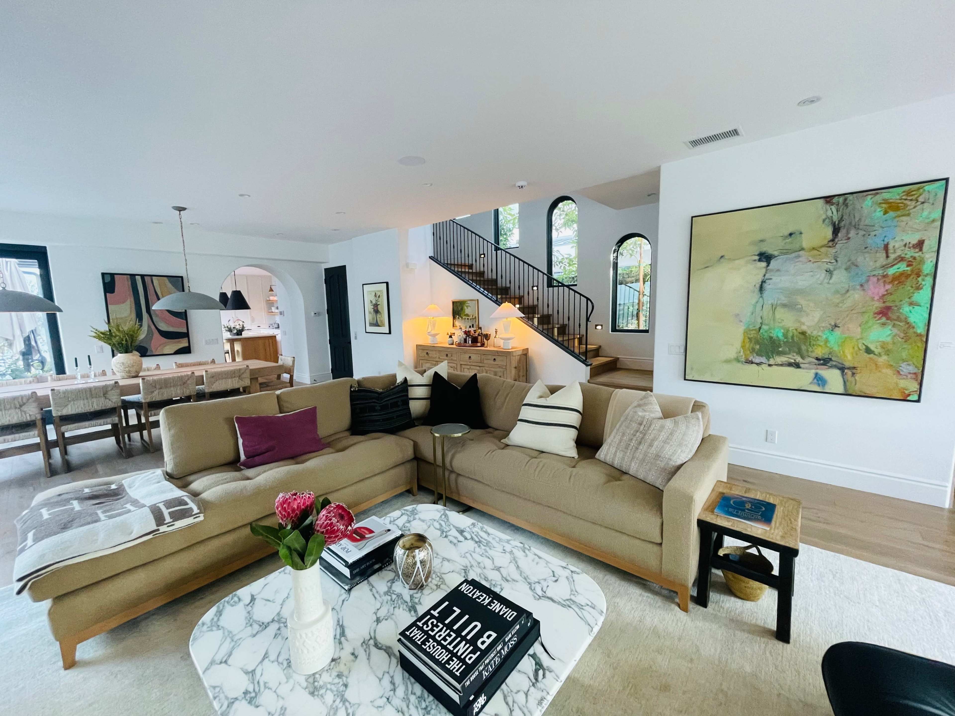 A modern living room features a large beige sectional sofa, a marble coffee table, and an abstract painting, with a dining area visible in the background.