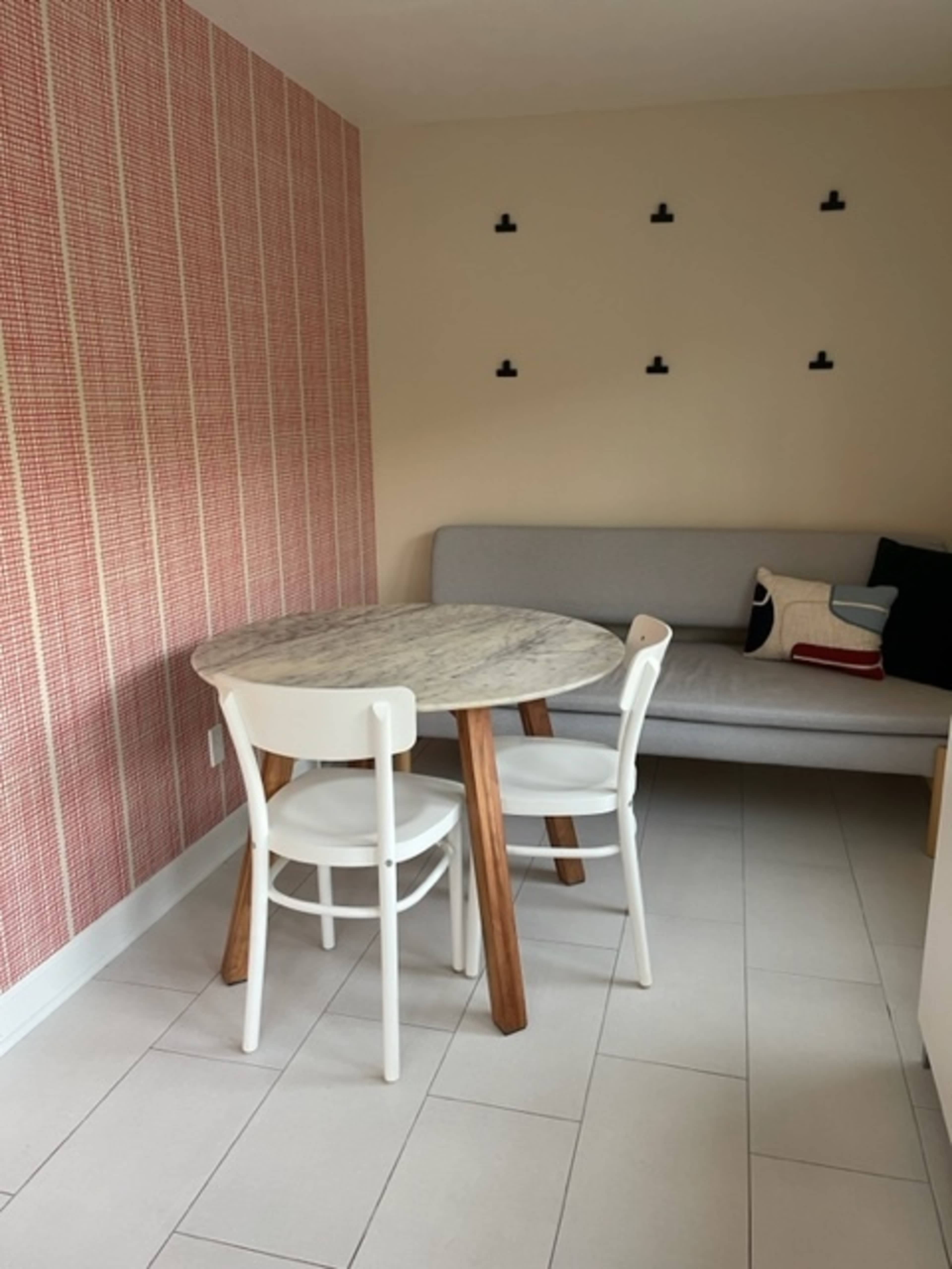 A small dining area features a round table with a marble top and two white chairs next to a gray sofa against a patterned red wall.