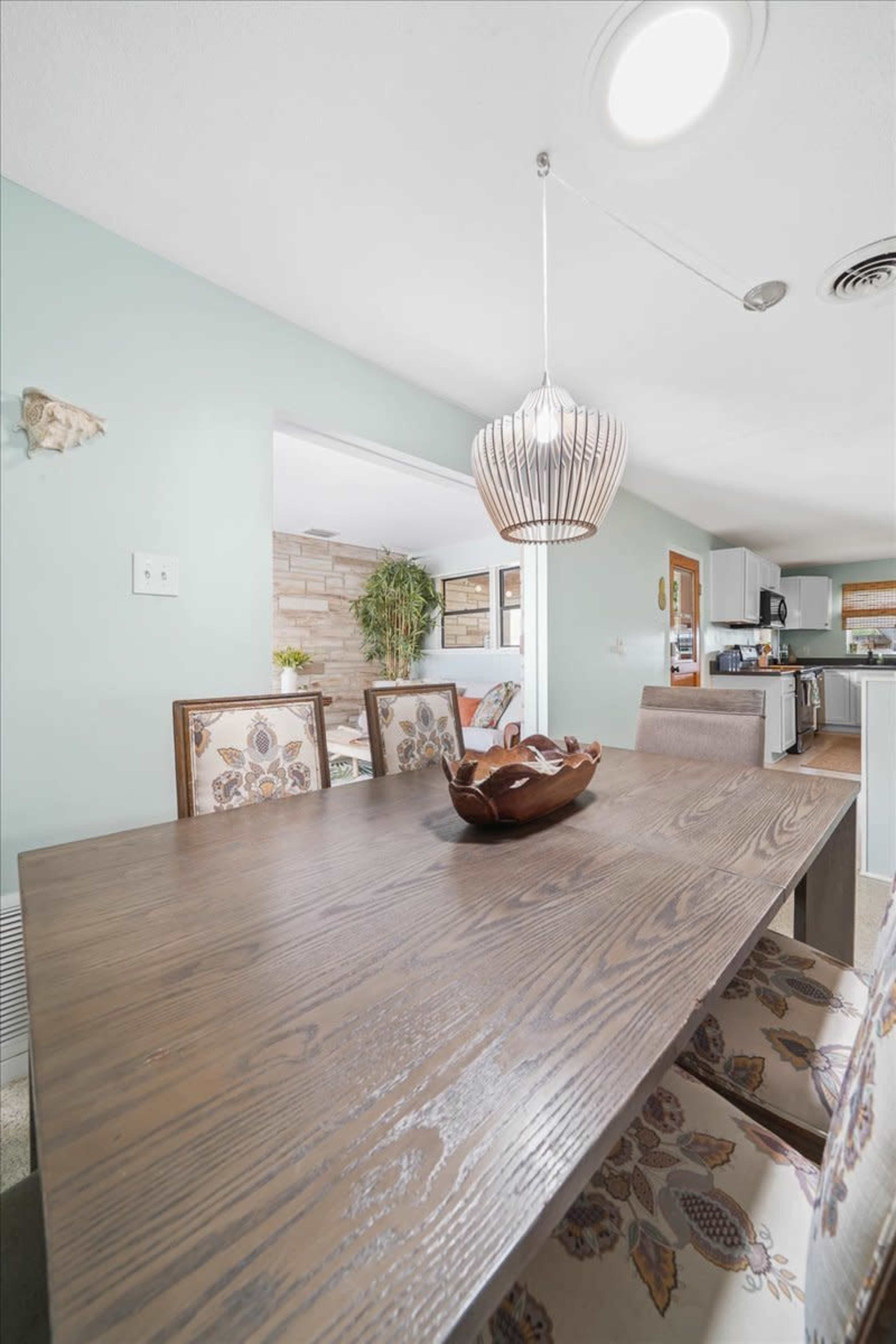 A wooden dining table is set in a light-colored room with a pendant light hanging above and a view of a kitchen in the background.