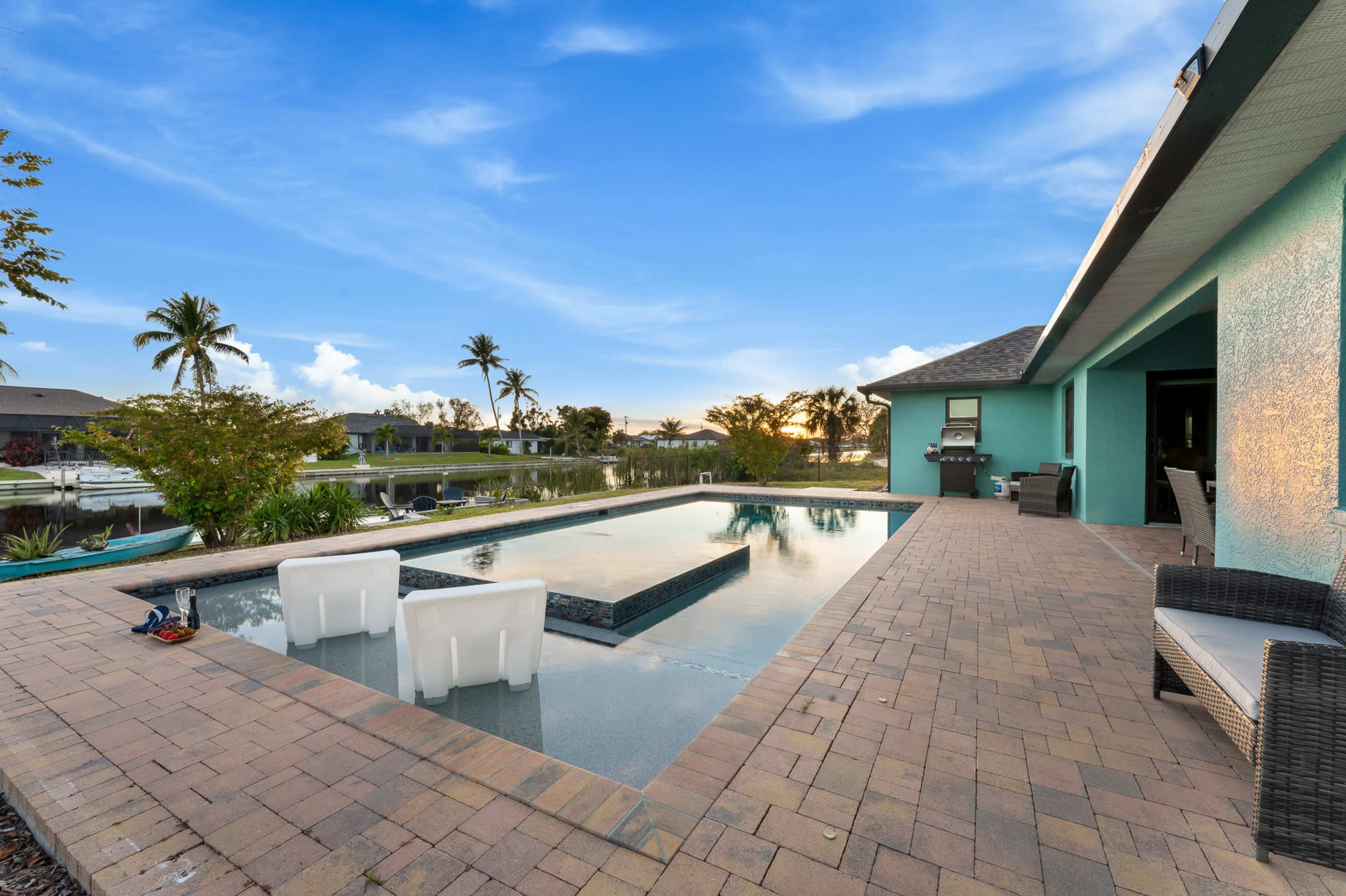 A backyard with a rectangular pool surrounded by paver stones, adjacent to a teal-colored house and palm trees in the background.
