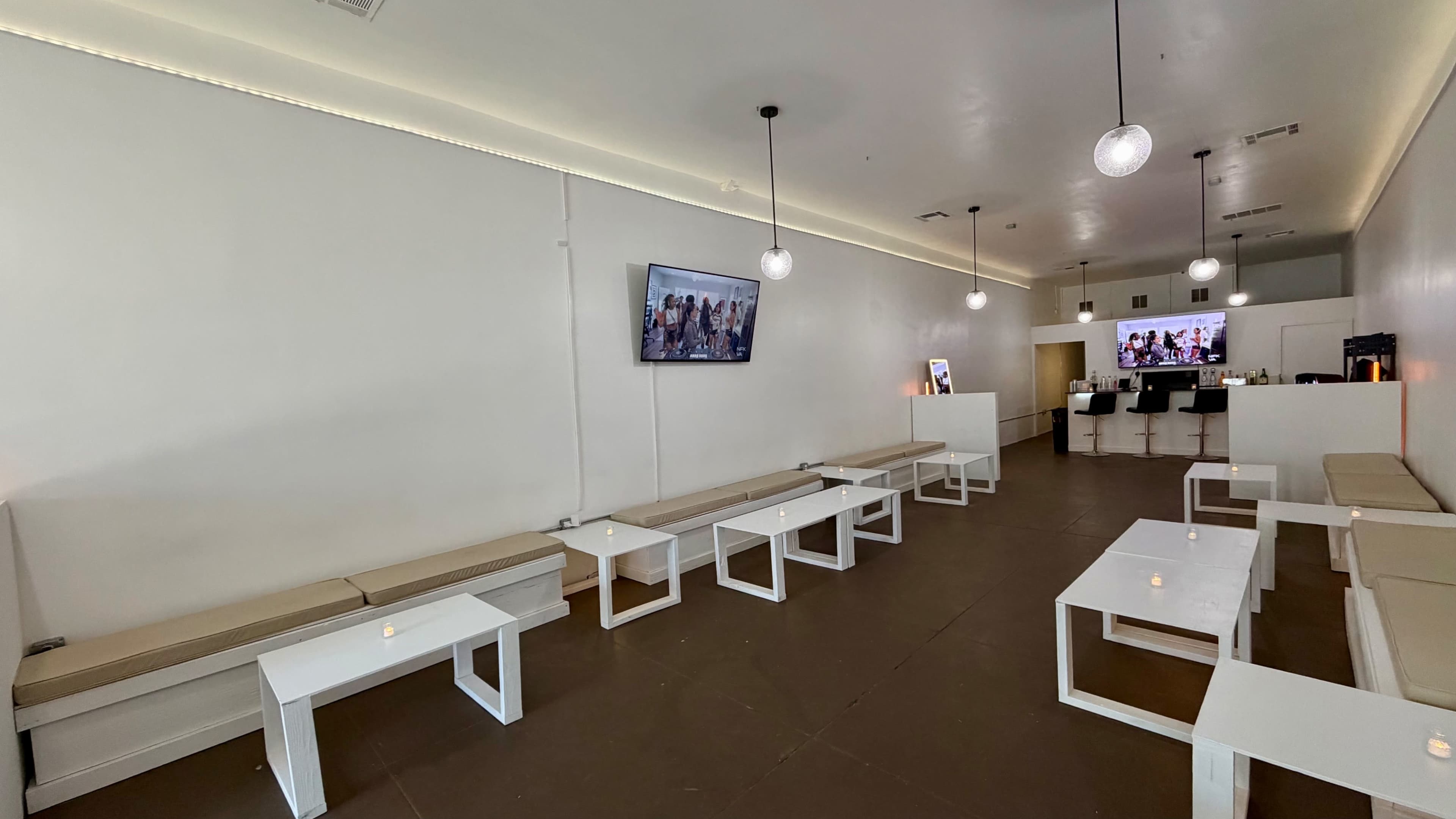 The image shows a modern, minimally furnished interior of a lounge or café with white walls, several low benches, small white tables, and a couple of screens displaying images.