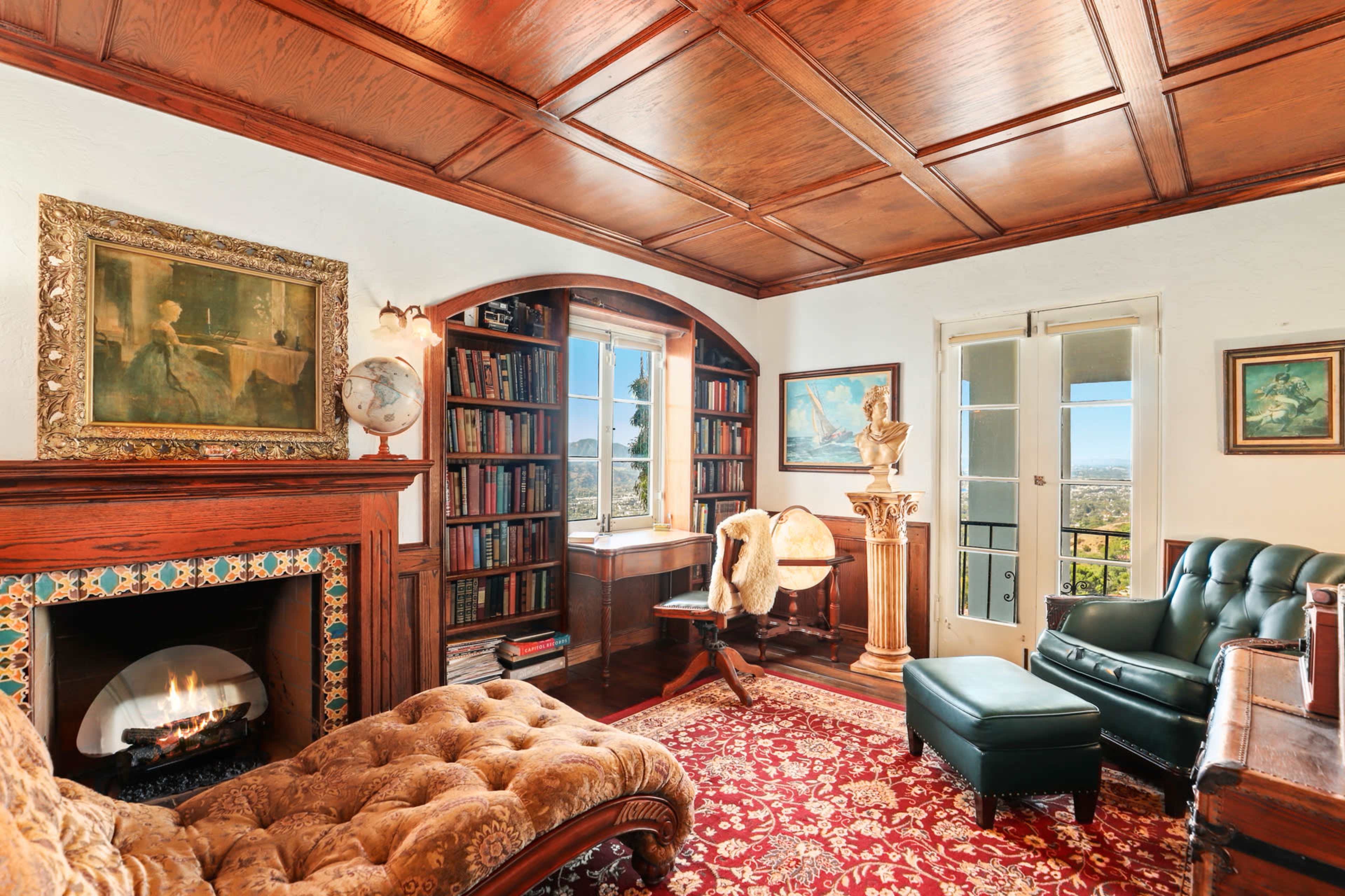 A cozy library with a wooden ceiling, a fireplace, bookshelves filled with books, and a view through a large window.