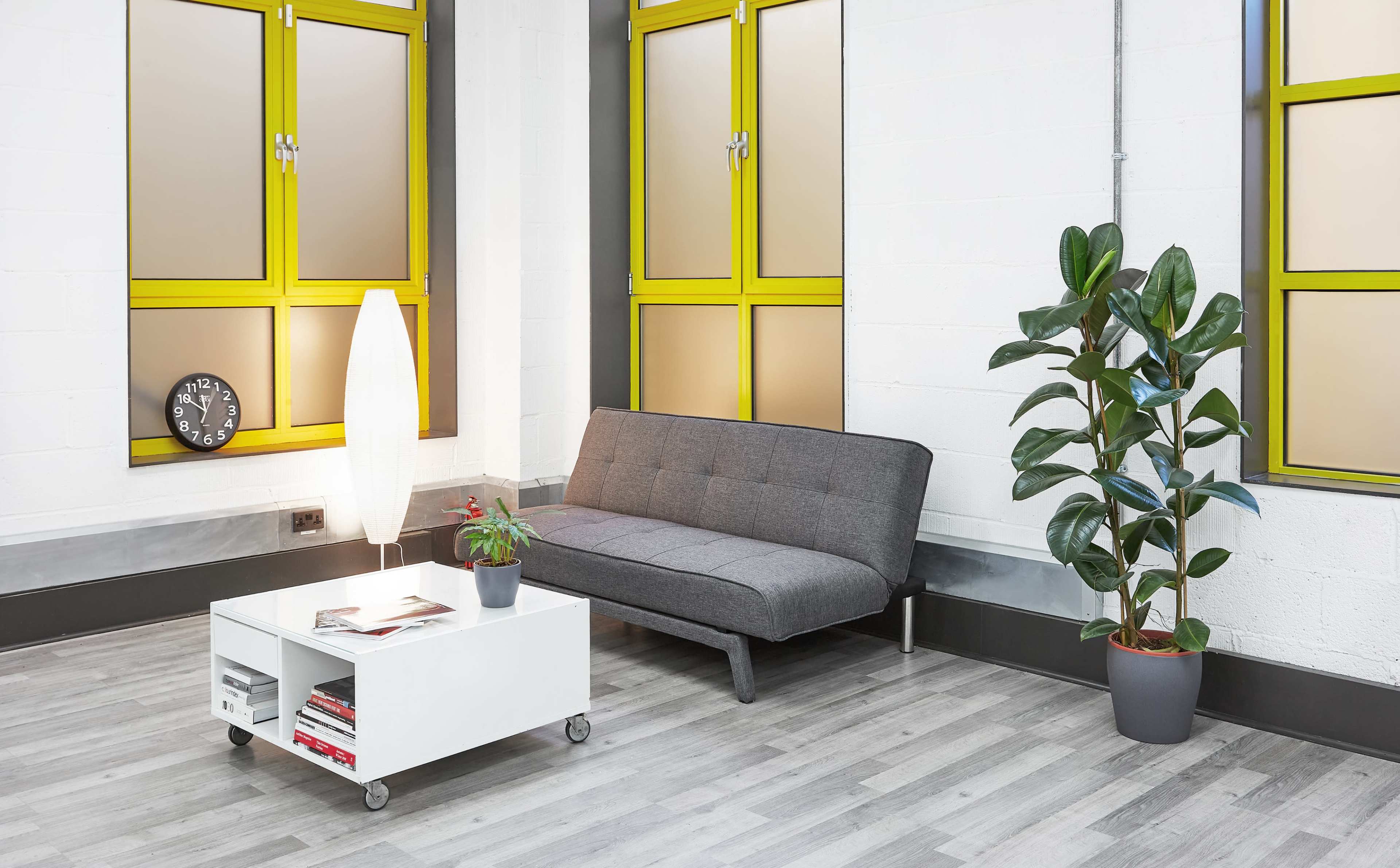 The interior features a gray sofa, a white coffee table on wheels, a plant, and large windows with yellow frames, all set in a brightly lit room.