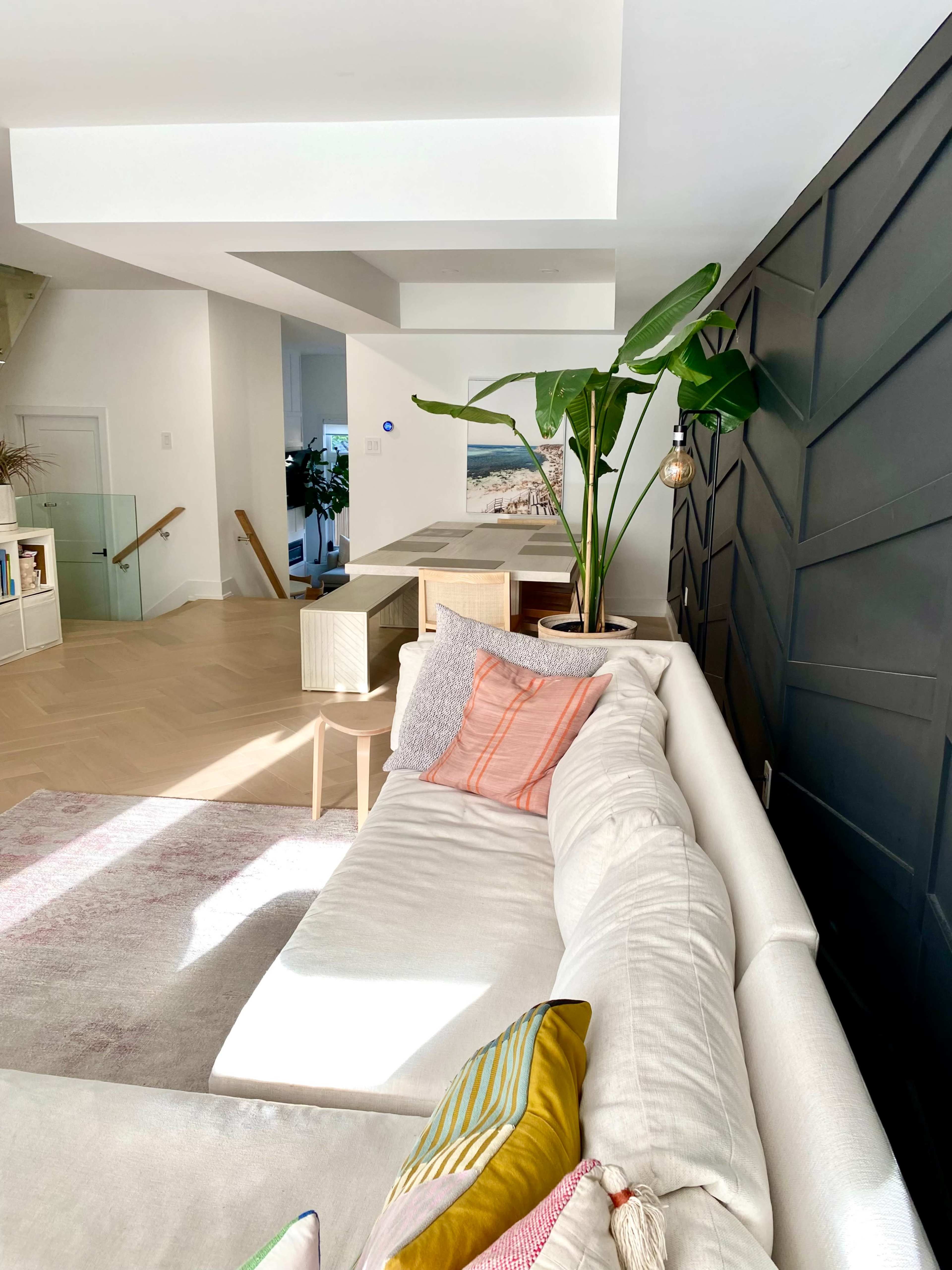 A white sectional sofa is positioned in a bright living space, adjacent to a dining area with a wooden table and potted plants.